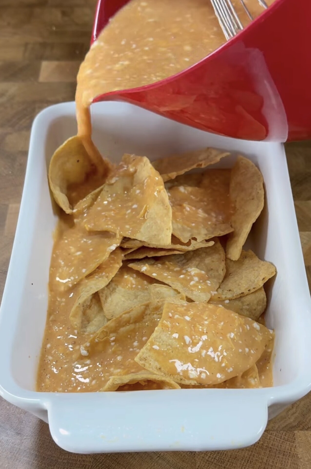 A red bowl is pouring a creamy orange sauce over tortilla chips in a white rectangular baking dish, preparing a cheesy nacho or casserole dish on a wooden surface.