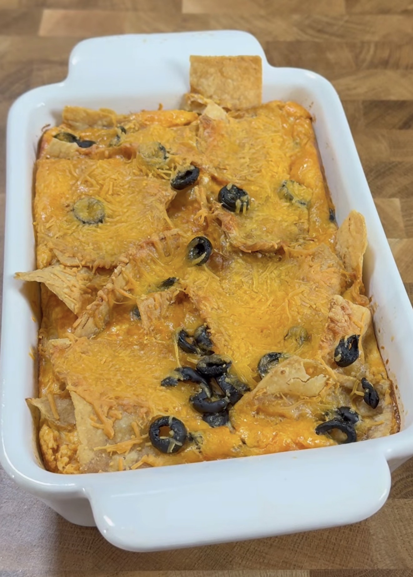 A white baking dish filled with a layered casserole topped with melted cheddar cheese, tortilla chips, and sliced black olives, set on a wooden surface.