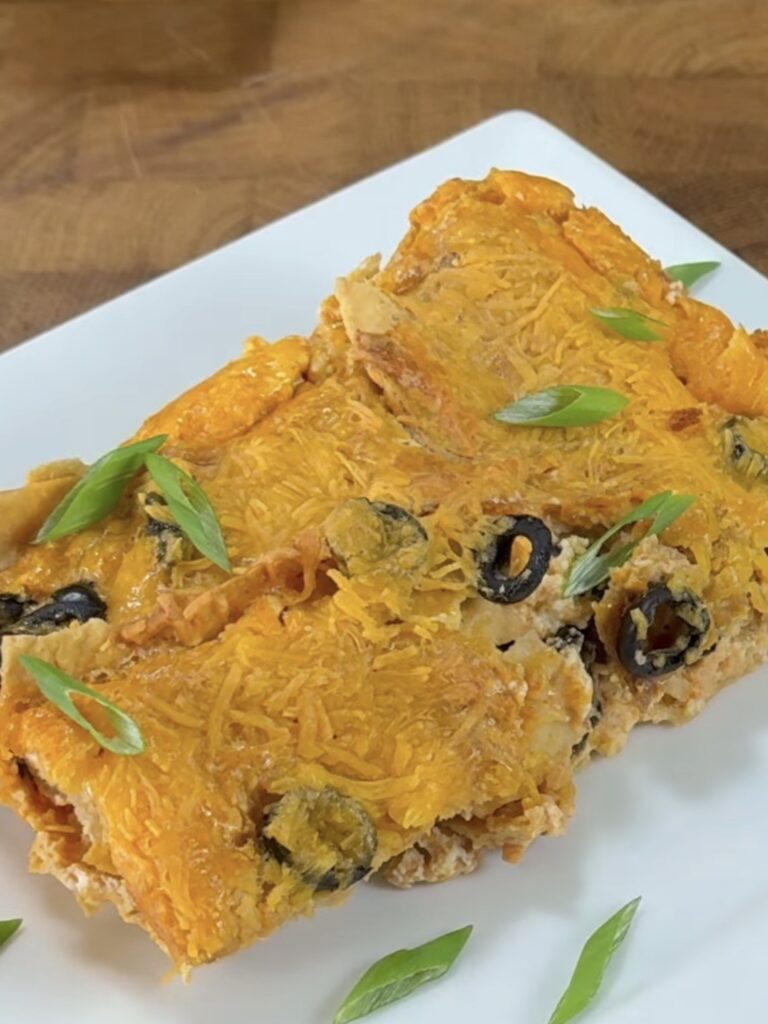 A close-up of cheesy enchiladas topped with sliced black olives and green onions, served on a white plate with a wooden background.