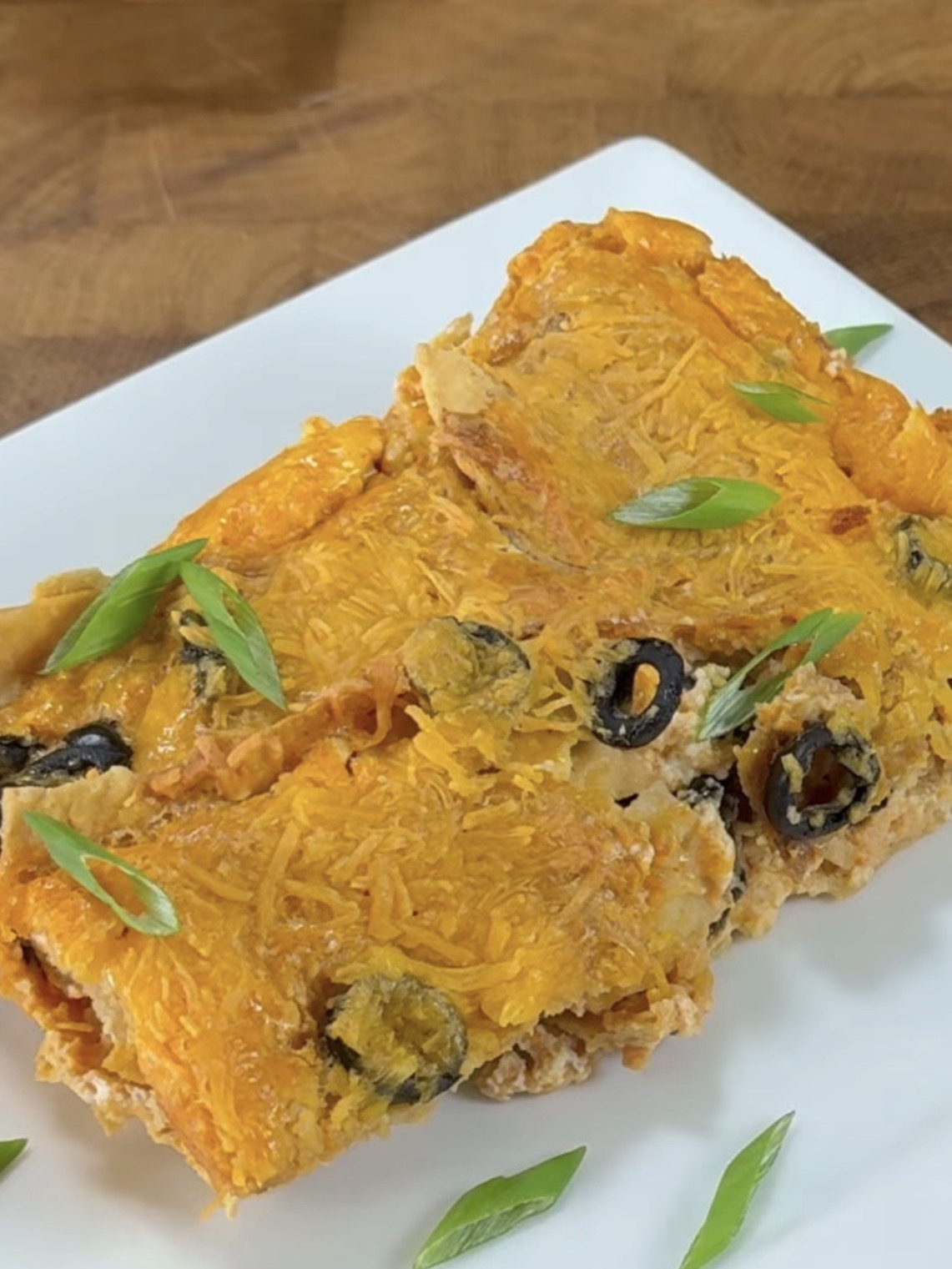 A close-up of cheesy enchiladas topped with sliced black olives and green onions, served on a white plate with a wooden background.