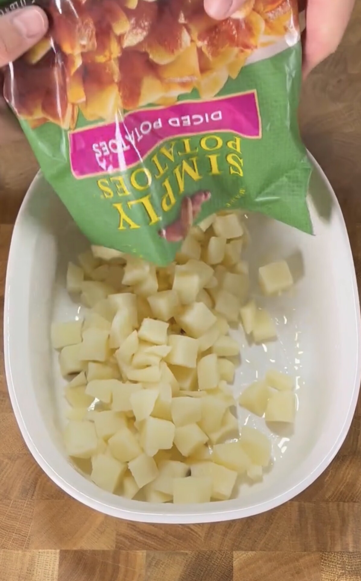 A bag of diced potatoes is being poured into a white baking dish on a wooden surface.