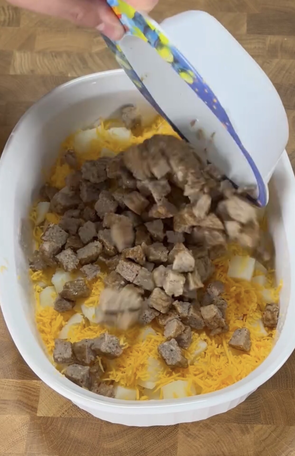 A hand is pouring cooked sausage pieces from a bowl into a white baking dish containing shredded cheese and diced potatoes on a wooden surface.