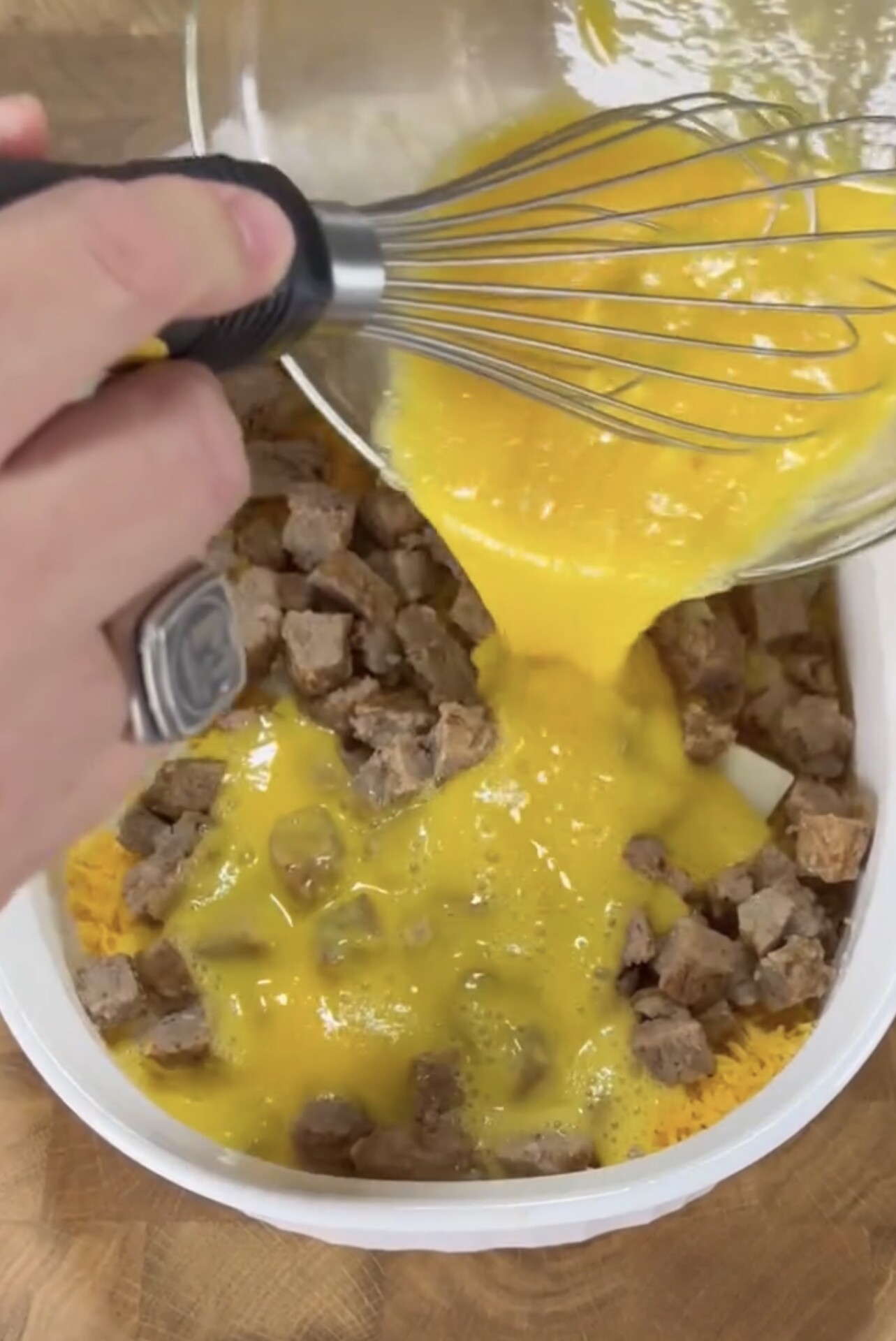 A hand holding a whisk pours beaten eggs from a glass bowl over a mixture of cooked sausage and shredded cheese in a white baking dish.