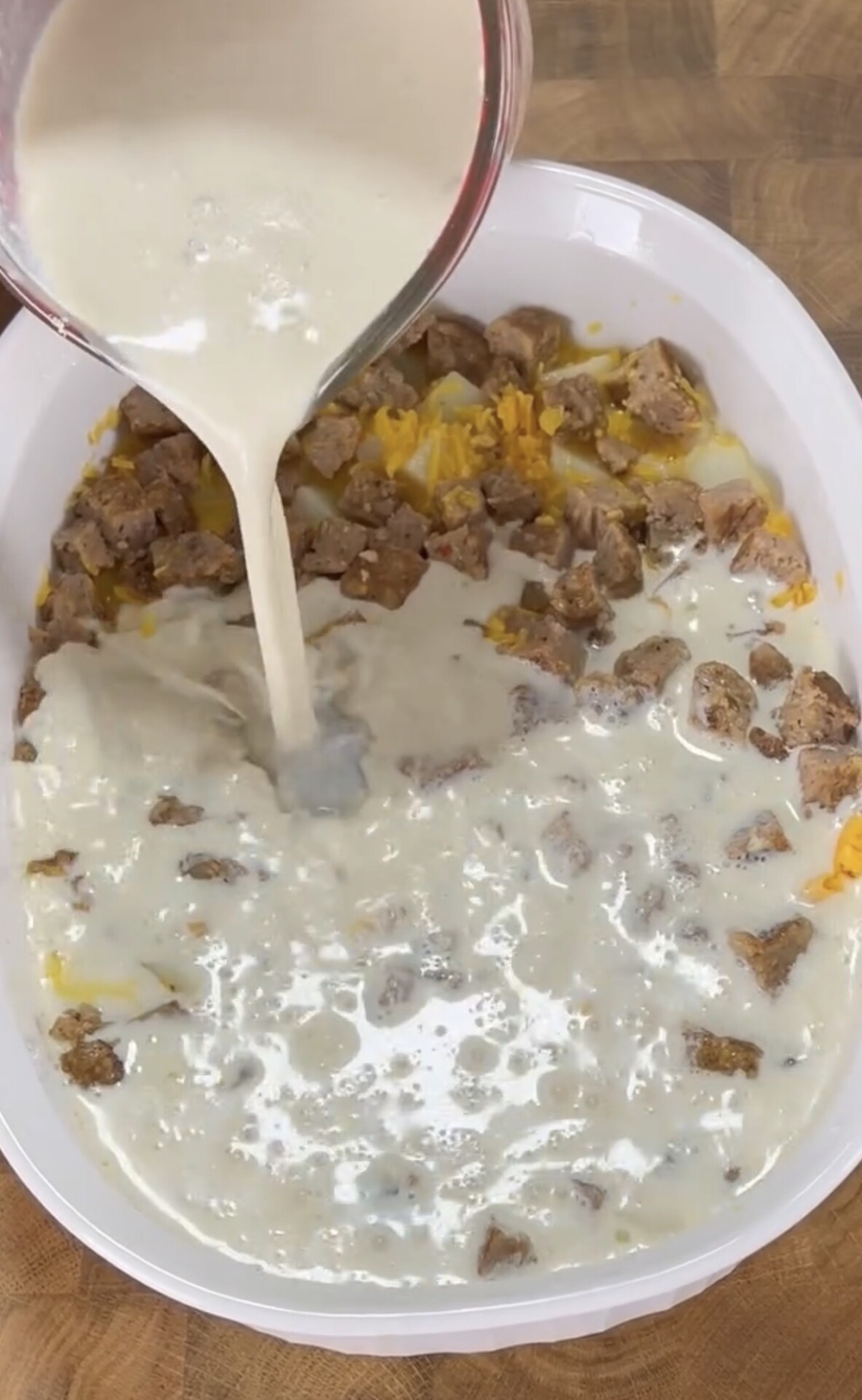 A white dish filled with chopped cooked sausage, shredded cheese, and potatoes is being covered with a creamy liquid mixture poured from a measuring cup. The dish sits on a wooden surface.