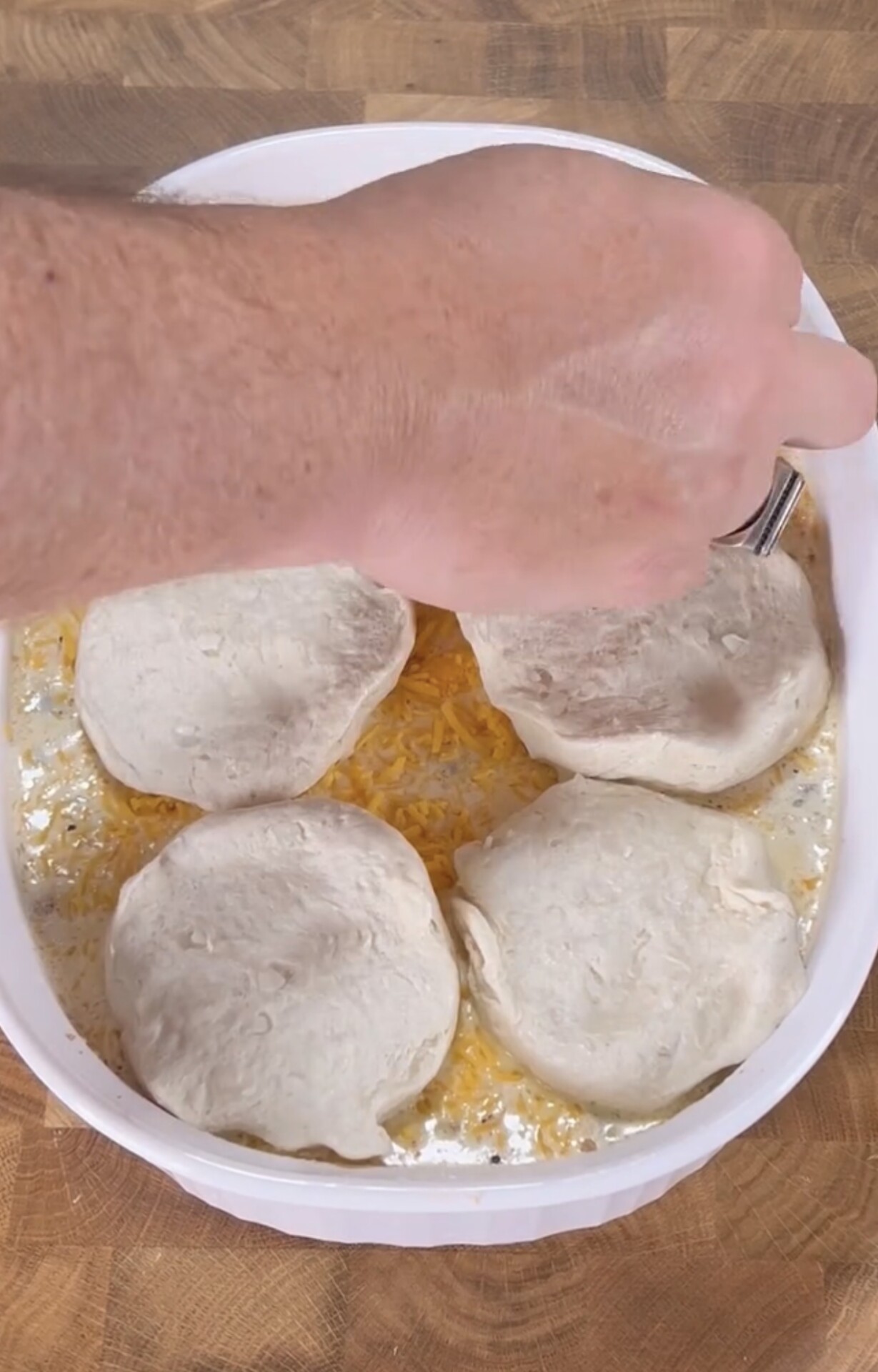 A hand sprinkles shredded cheese over four raw biscuit dough rounds in a white baking dish filled with creamy sauce on a wooden surface.