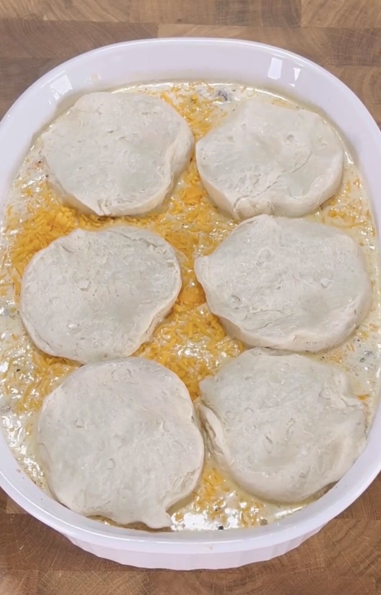 A white baking dish filled with a creamy mixture topped with shredded cheese and six unbaked biscuit dough rounds, ready to be baked.