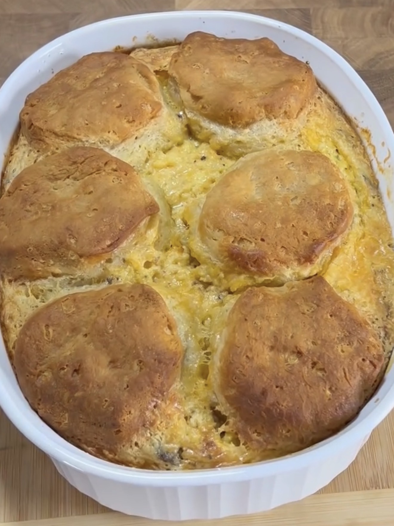 A baked casserole topped with six golden-brown biscuits, set in a white oval dish on a wooden surface. The casserole appears creamy and cheesy underneath the biscuits.