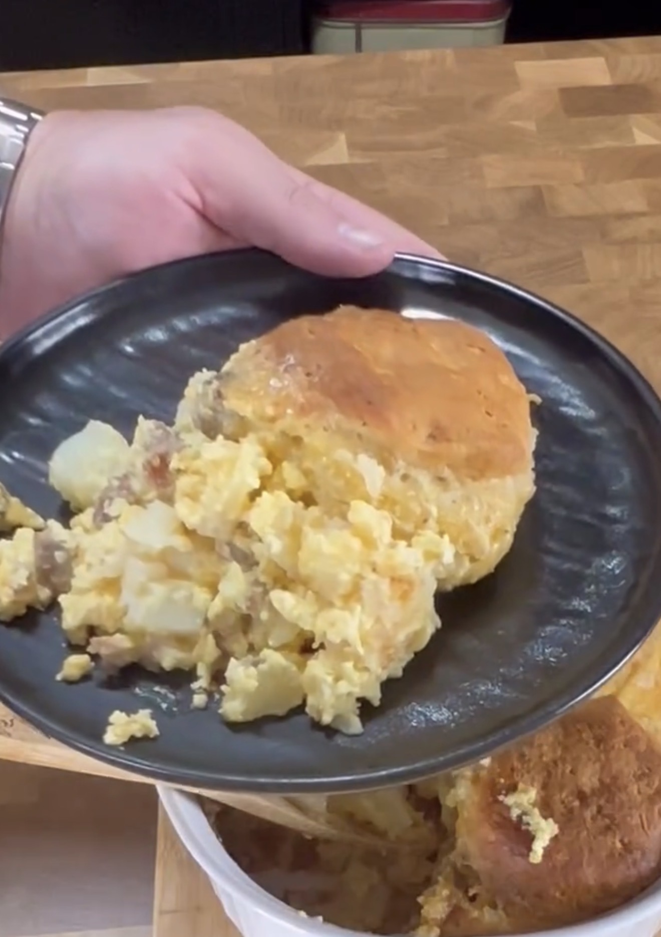 A hand holds a black plate containing a serving of a baked dish with scrambled eggs, potatoes, sausage, and a golden-brown biscuit topping. The plate is above a pan with the same casserole.