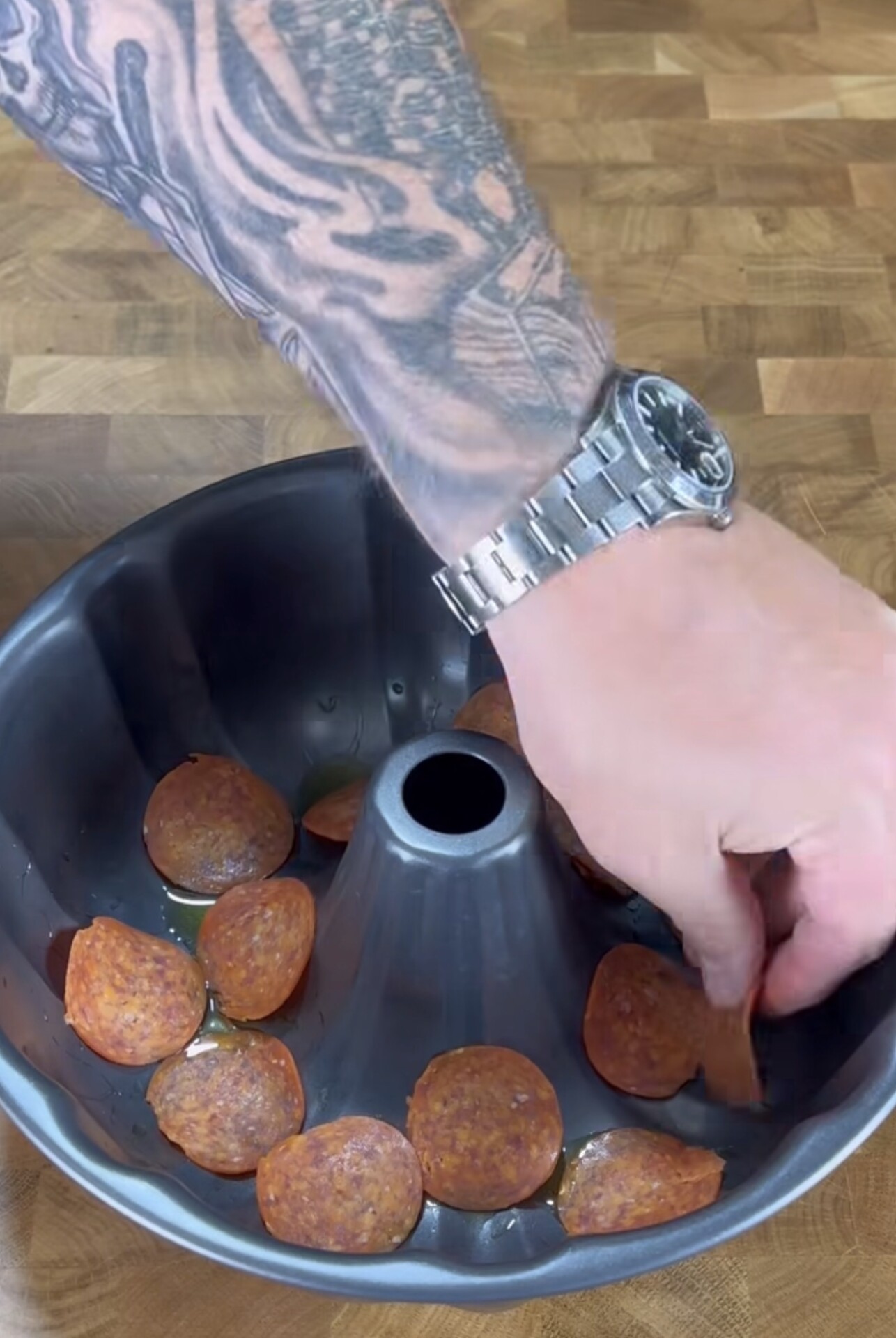 A tattooed arm with a watch places slices of pepperoni into a greased bundt cake pan on a wooden countertop.