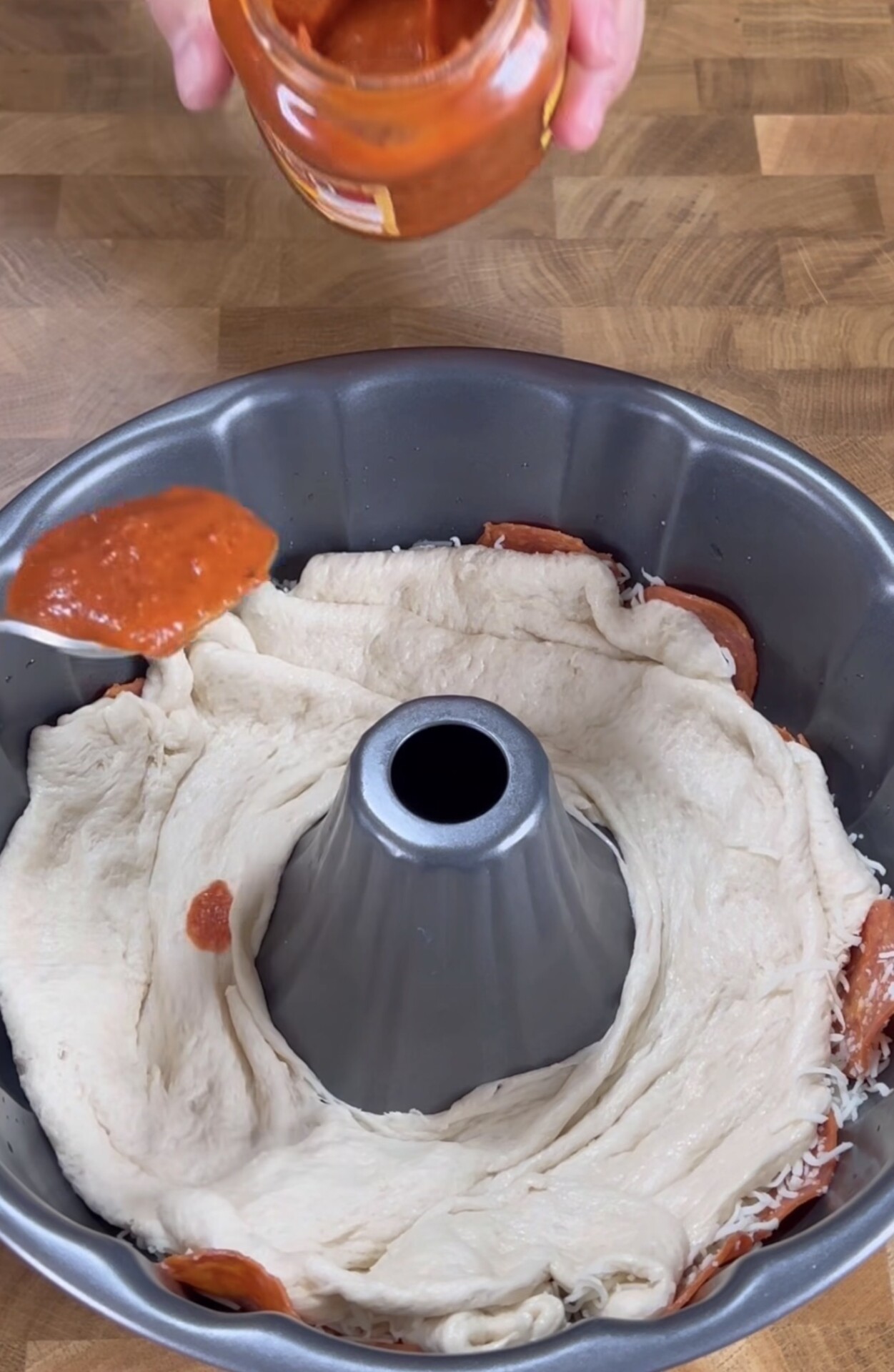 A bundt pan lined with pizza dough, cheese, and pepperoni, with a hand above it spooning tomato sauce from a jar onto the dough. The pan sits on a wooden surface.