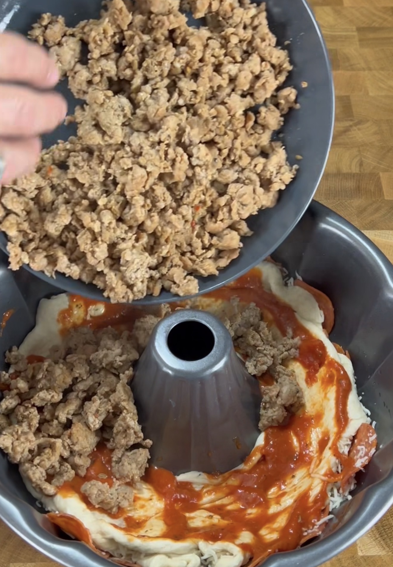 A hand pours crumbled cooked sausage into a bundt pan layered with dough, tomato sauce, and more sausage, on a wooden countertop.