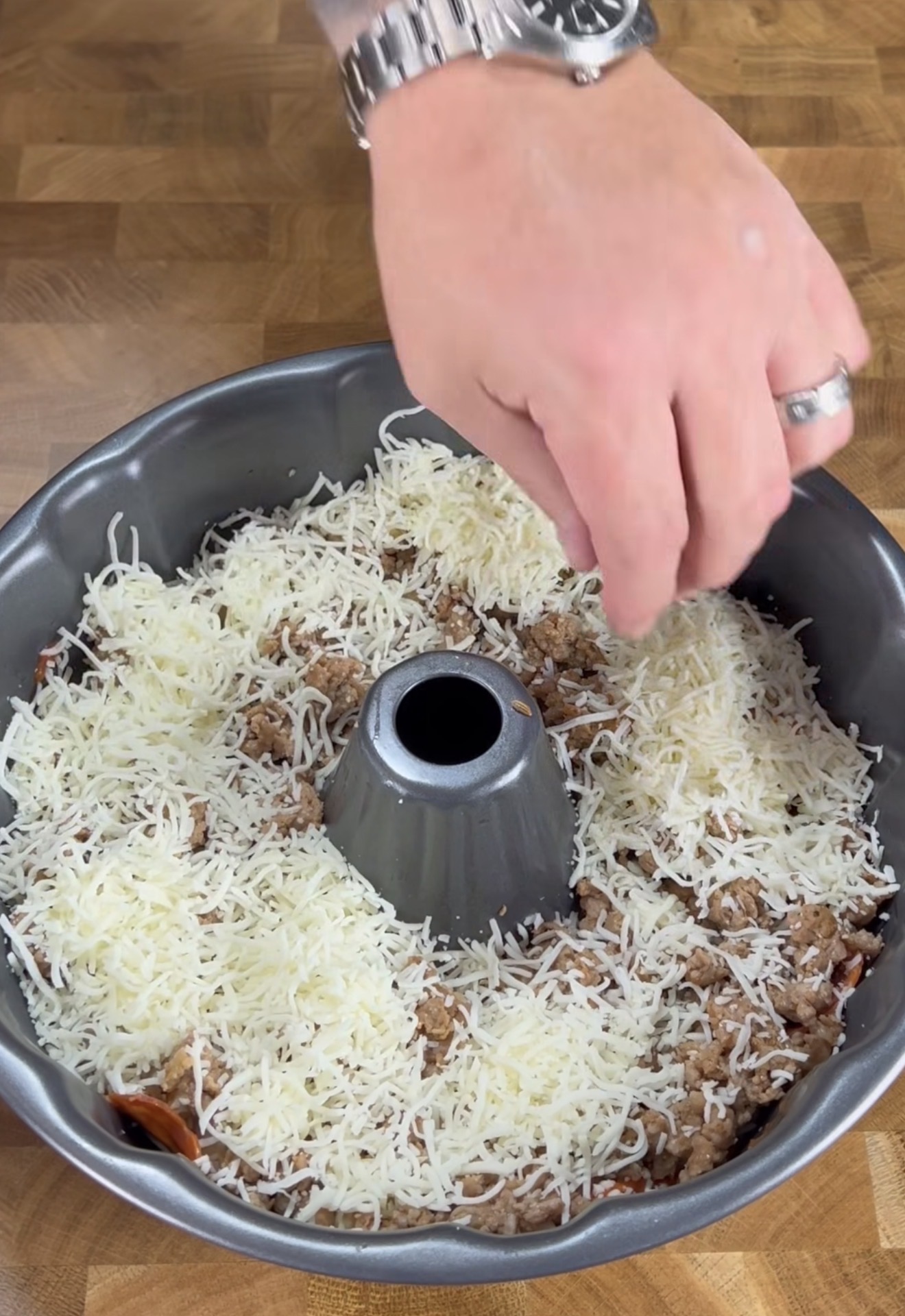 A hand sprinkles shredded cheese over cooked ground meat in a bundt pan on a wooden surface.