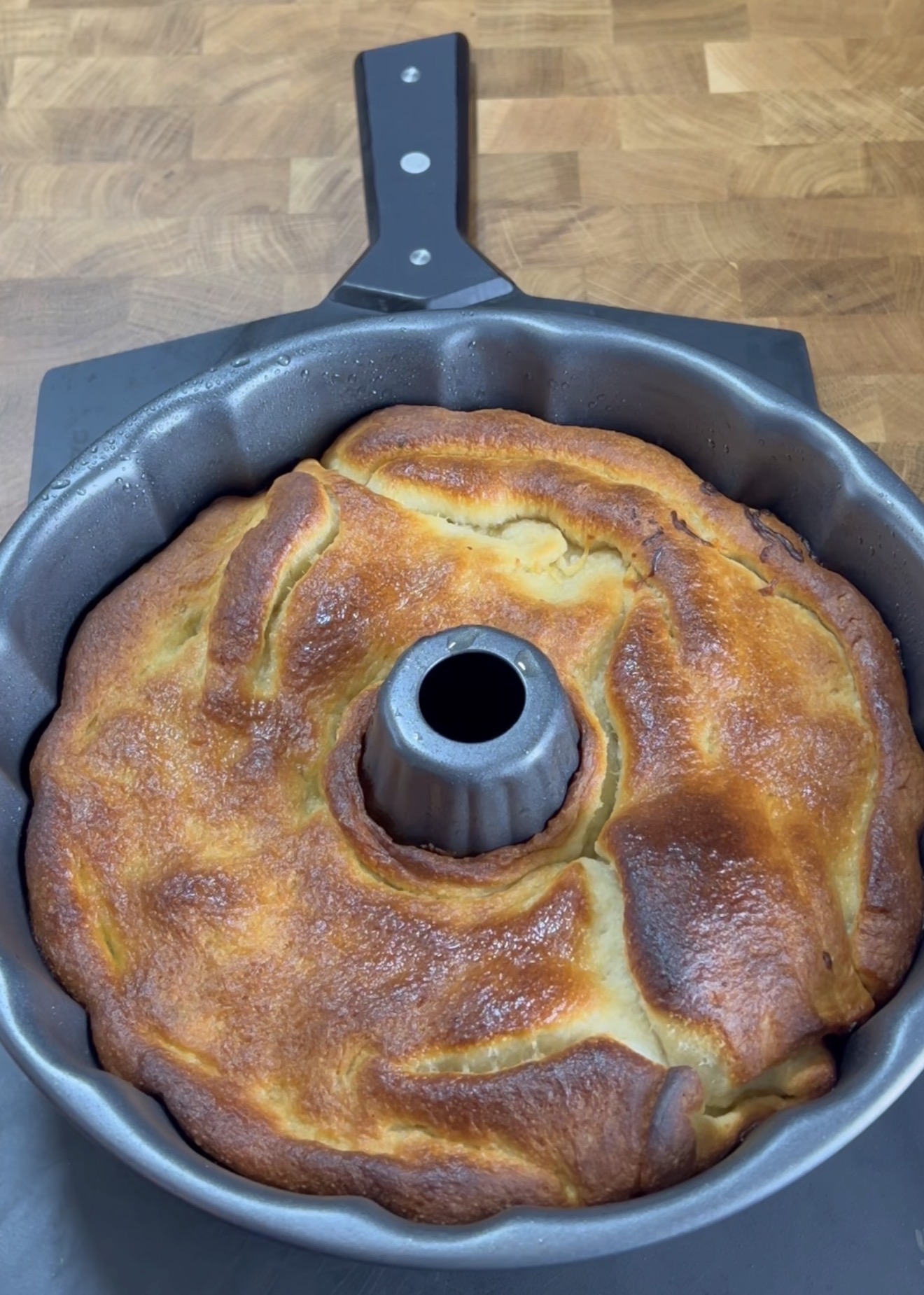 A golden-brown baked cake with a slightly uneven surface sits in a metal Bundt pan on a wooden countertop. The cake has risen around the center tube of the pan.
