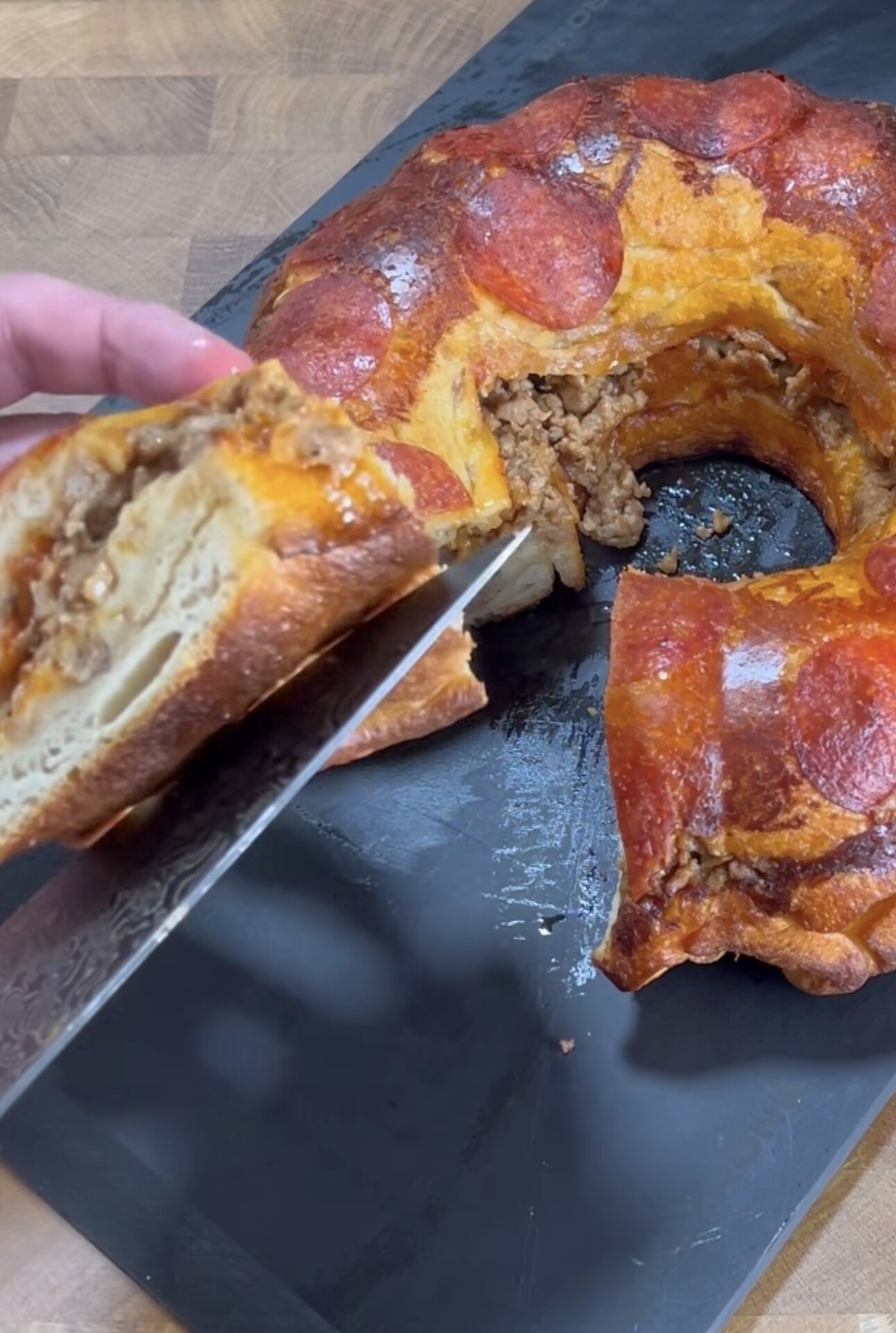 A hand slices into a bundt-shaped pizza ring topped with pepperoni, revealing a filling of ground meat and cheese, all placed on a black cutting board.