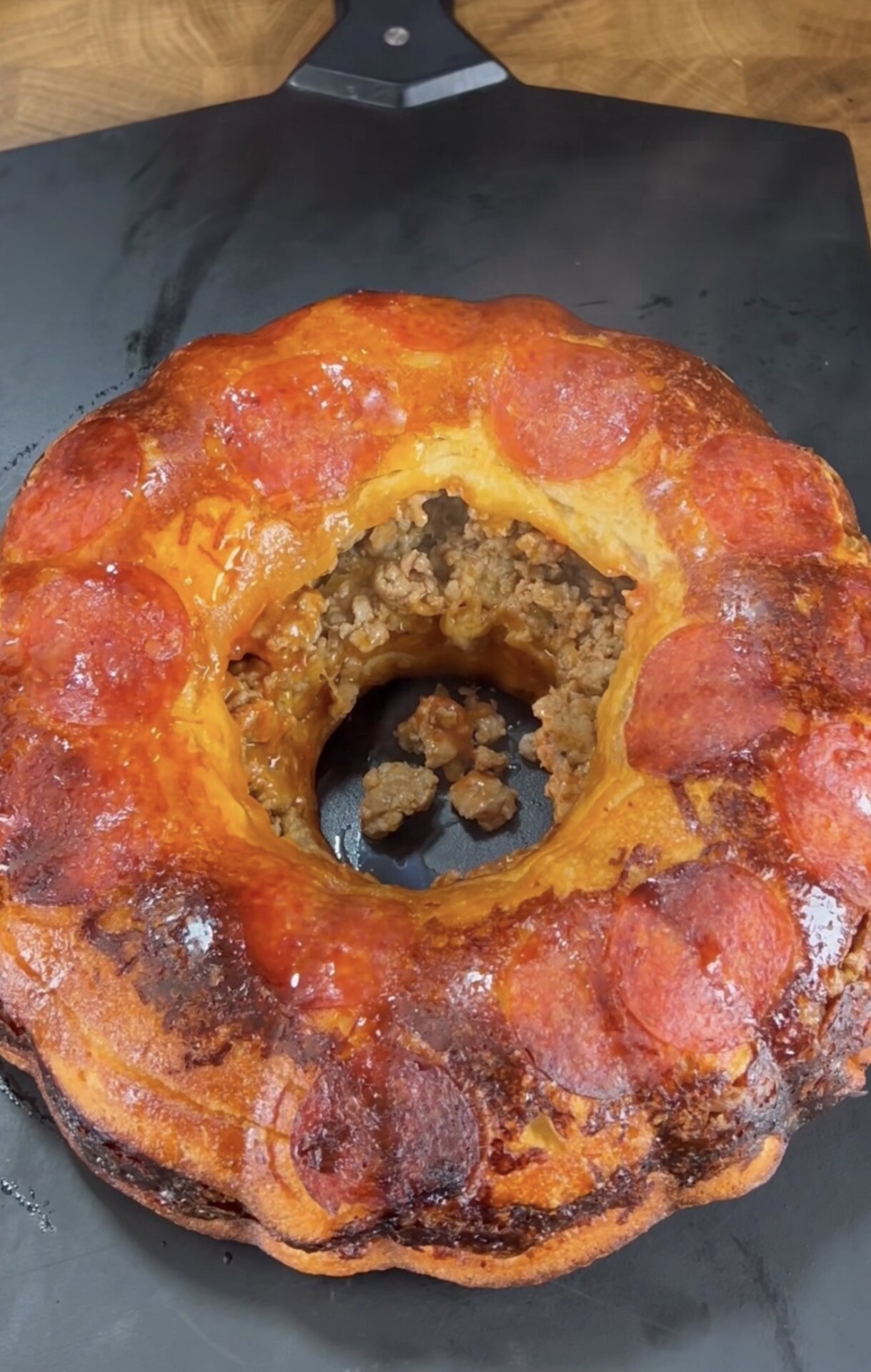 A round, golden-brown baked ring filled with cooked ground meat and topped with pepperoni slices, resting on a dark surface. A section is cut out, revealing the savory filling inside.