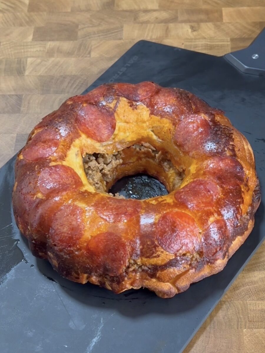 A golden-brown bundt-shaped bread loaf topped with pepperoni slices, with a filling visible inside, sits on a black cutting board on a wooden surface.