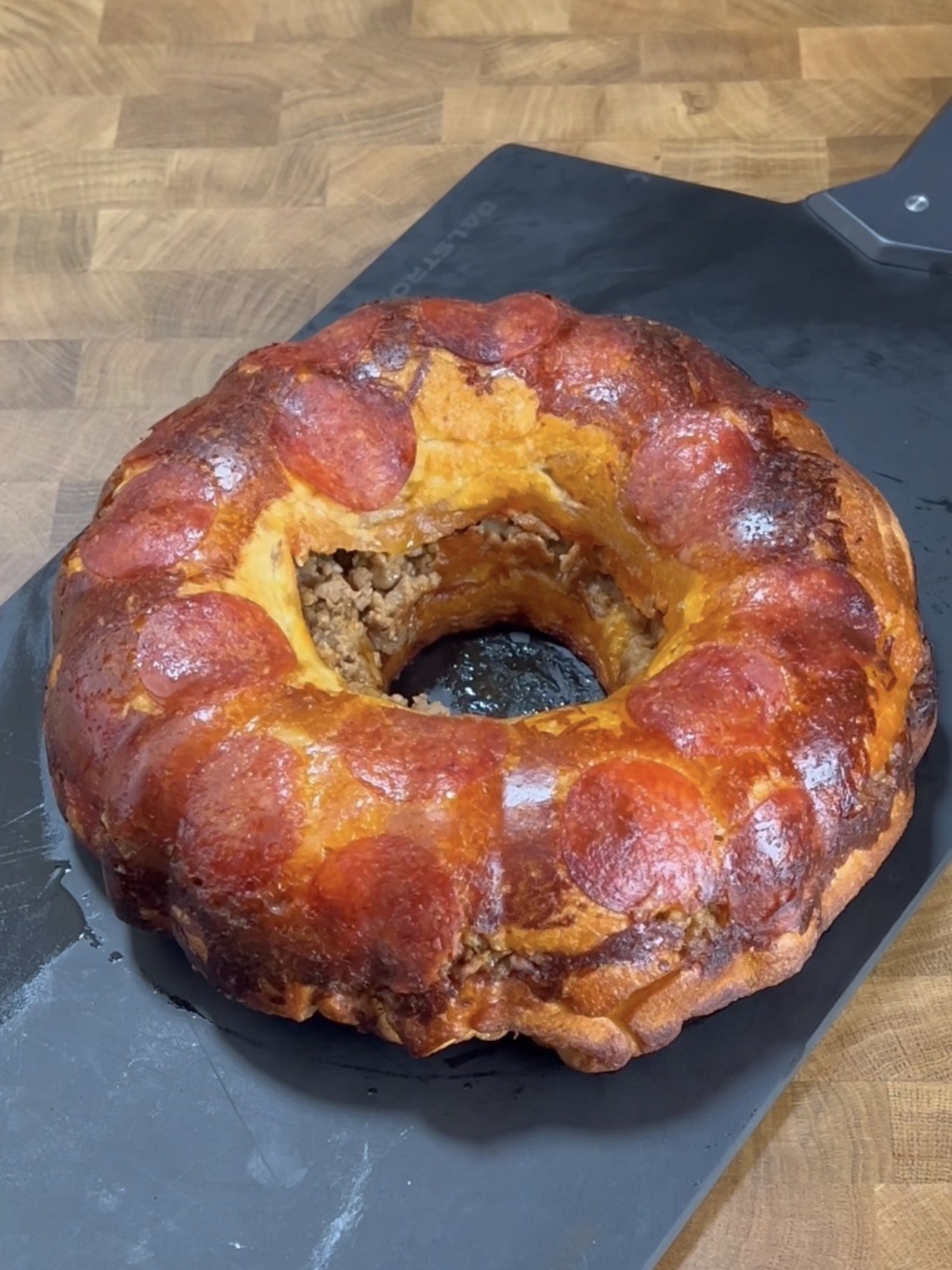 A golden-brown bundt-shaped bread loaf topped with pepperoni slices, with a filling visible inside, sits on a black cutting board on a wooden surface.