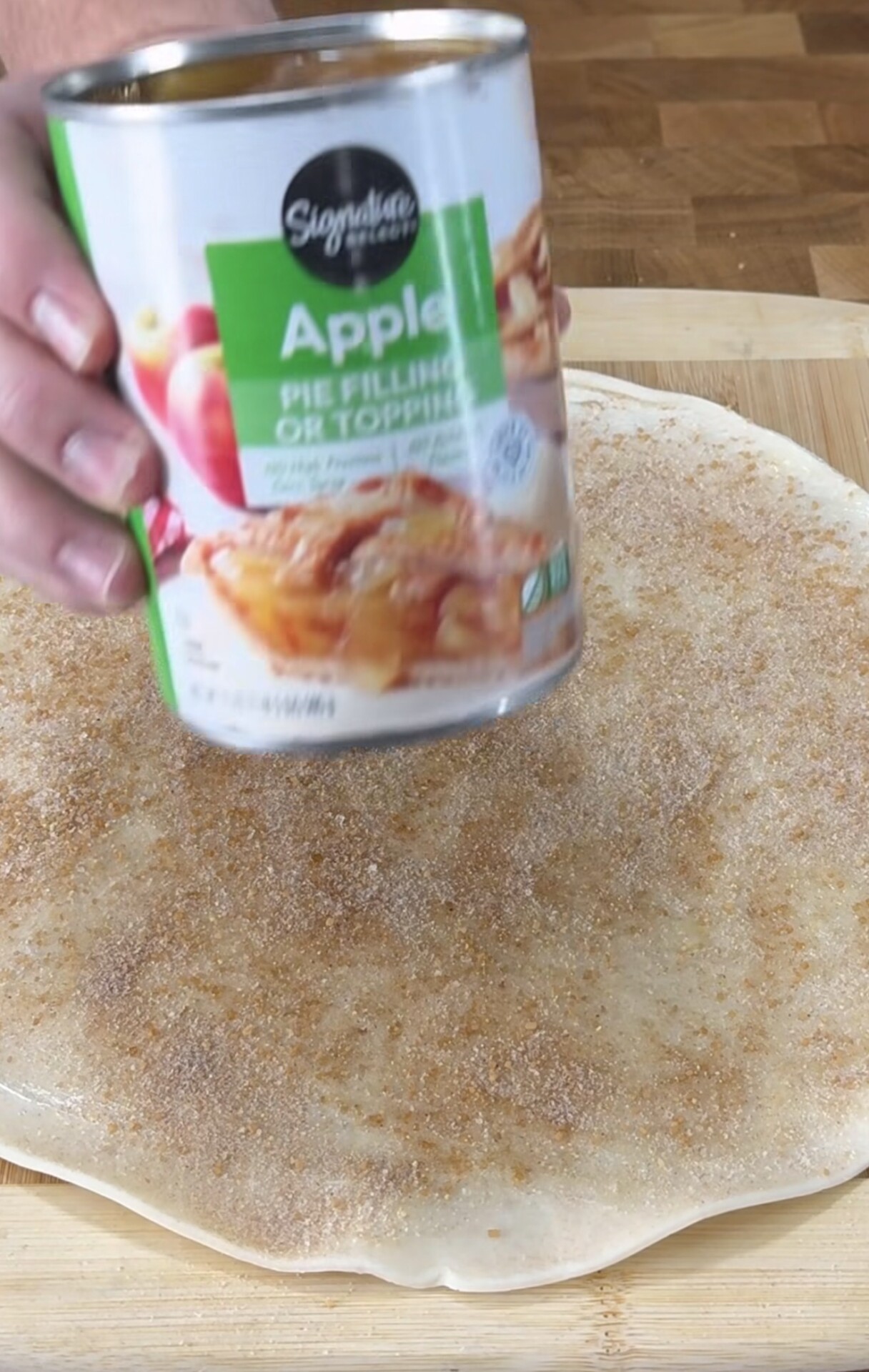 A hand holds a can of apple pie filling above rolled-out dough sprinkled with cinnamon sugar on a wooden surface.