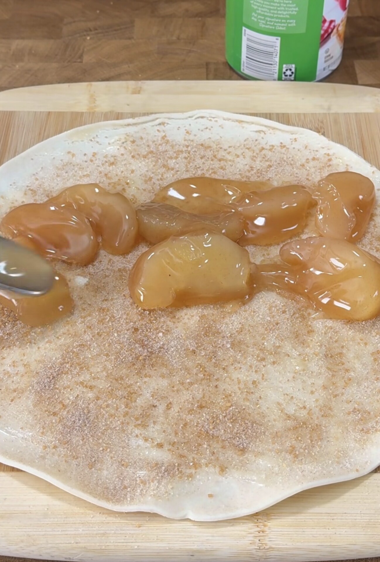 A tortilla is spread with cinnamon sugar and topped with spoonfuls of apple pie filling on a wooden cutting board, with a can and countertop in the background.
