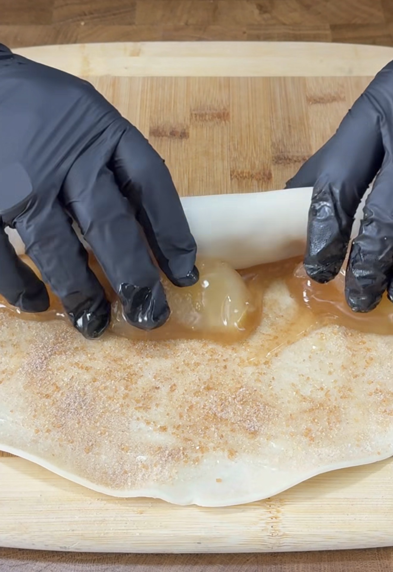 Hands in black gloves are rolling up a piece of dough filled with apple pie filling and sprinkled with cinnamon sugar on a wooden cutting board.