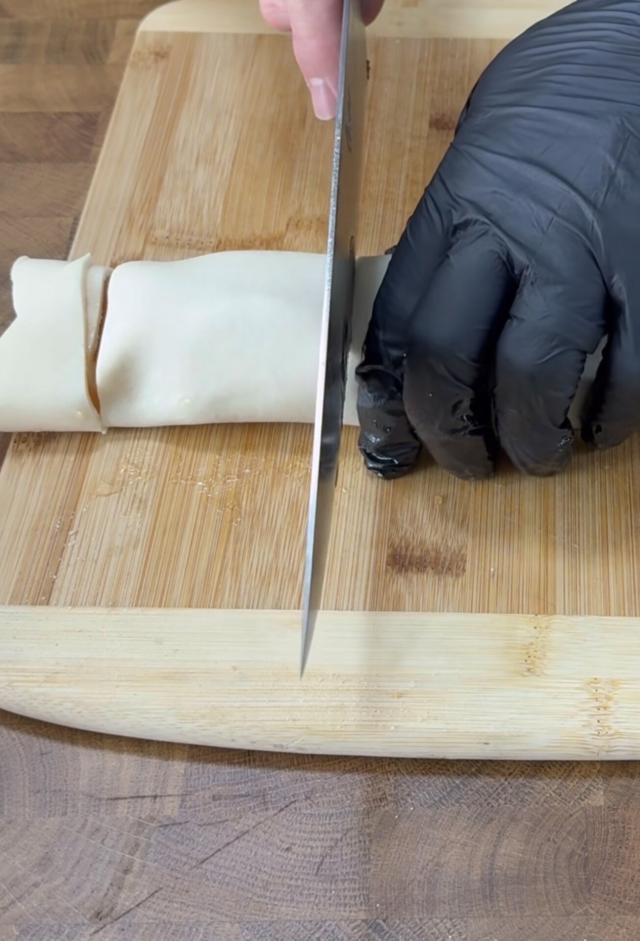 A hand in a black glove is slicing rolled dough on a wooden cutting board with a large knife.