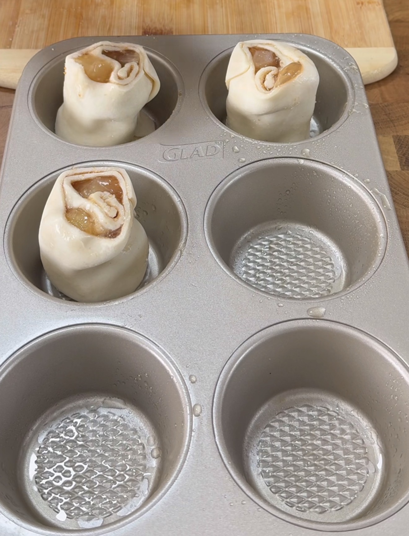 A muffin tin holding three unbaked apple pie rolls made of pastry dough with visible apple filling, while three other muffin cups remain empty and lightly greased. A wooden board is seen in the background.