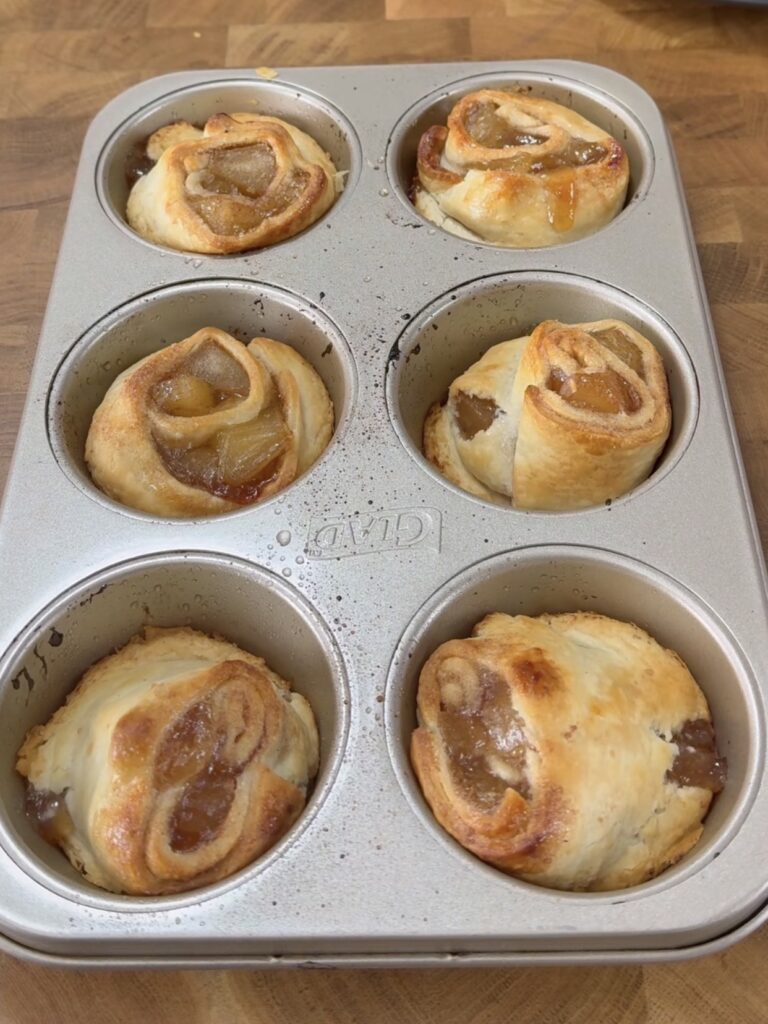 Six golden-brown puff pastry apple rose tarts are baked in a muffin tin and placed on a wooden surface. The pastries have visible swirls and apple filling peeking out from the flaky layers.