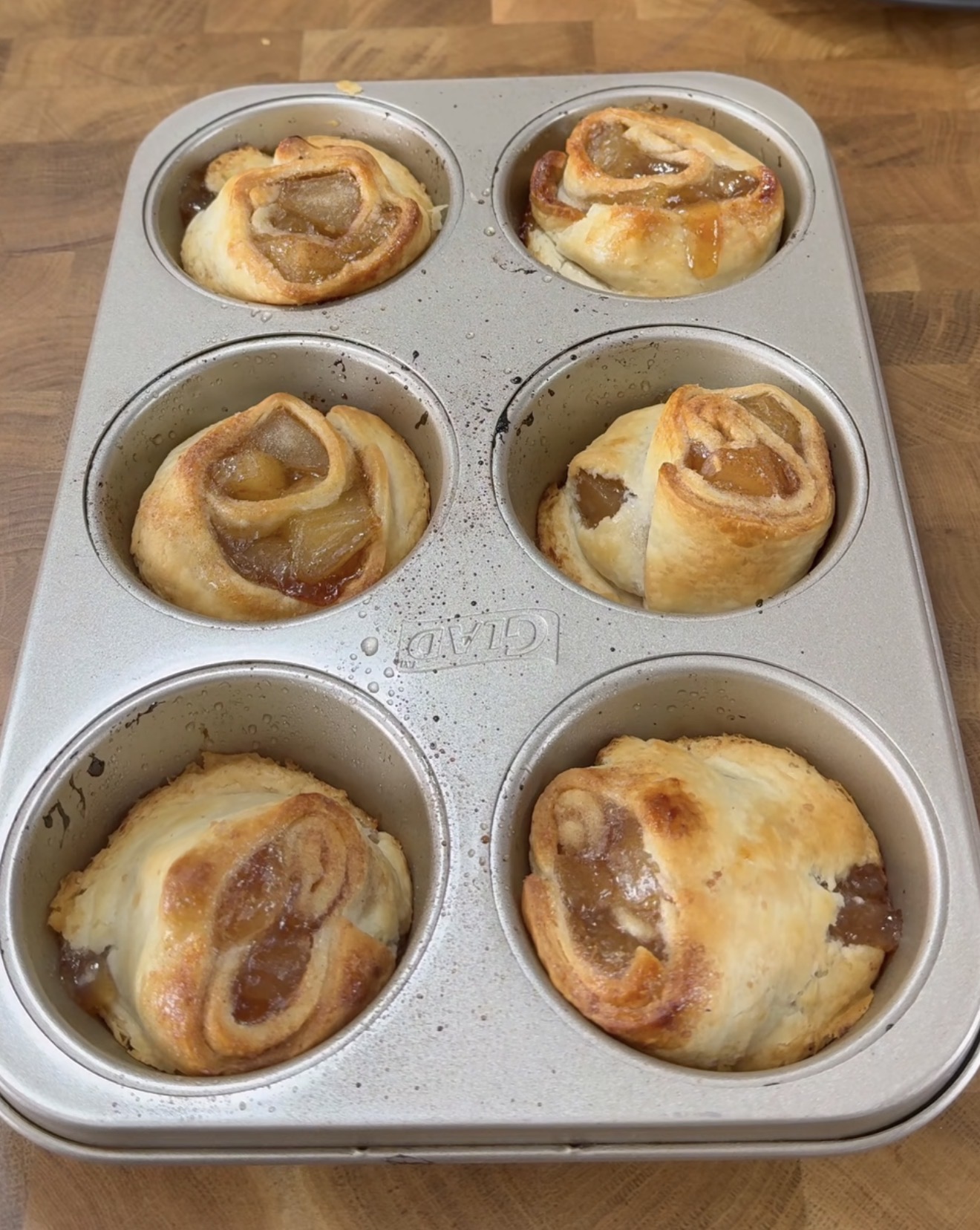 A metal muffin tin sits on a wooden surface, holding six baked pastry rolls with a golden-brown crust and swirled filling. Some rolls have a slight glaze and visible fruit or jam inside.