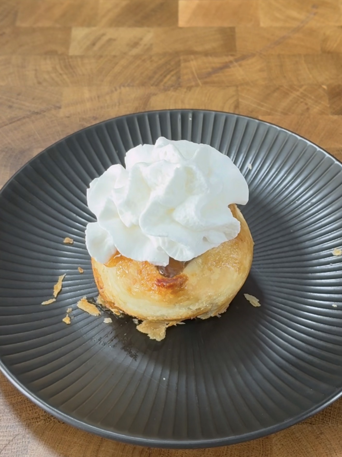 A round pastry topped with a swirl of whipped cream sits on a textured black plate, with a few flaky crumbs scattered around, placed on a wooden surface.