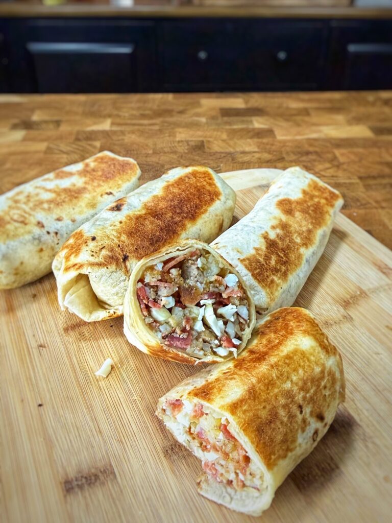 Four grilled burritos rest on a wooden cutting board, with one burrito cut open to reveal a filling of rice, meat, beans, chopped vegetables, and cheese. The background is a kitchen countertop and cabinets.