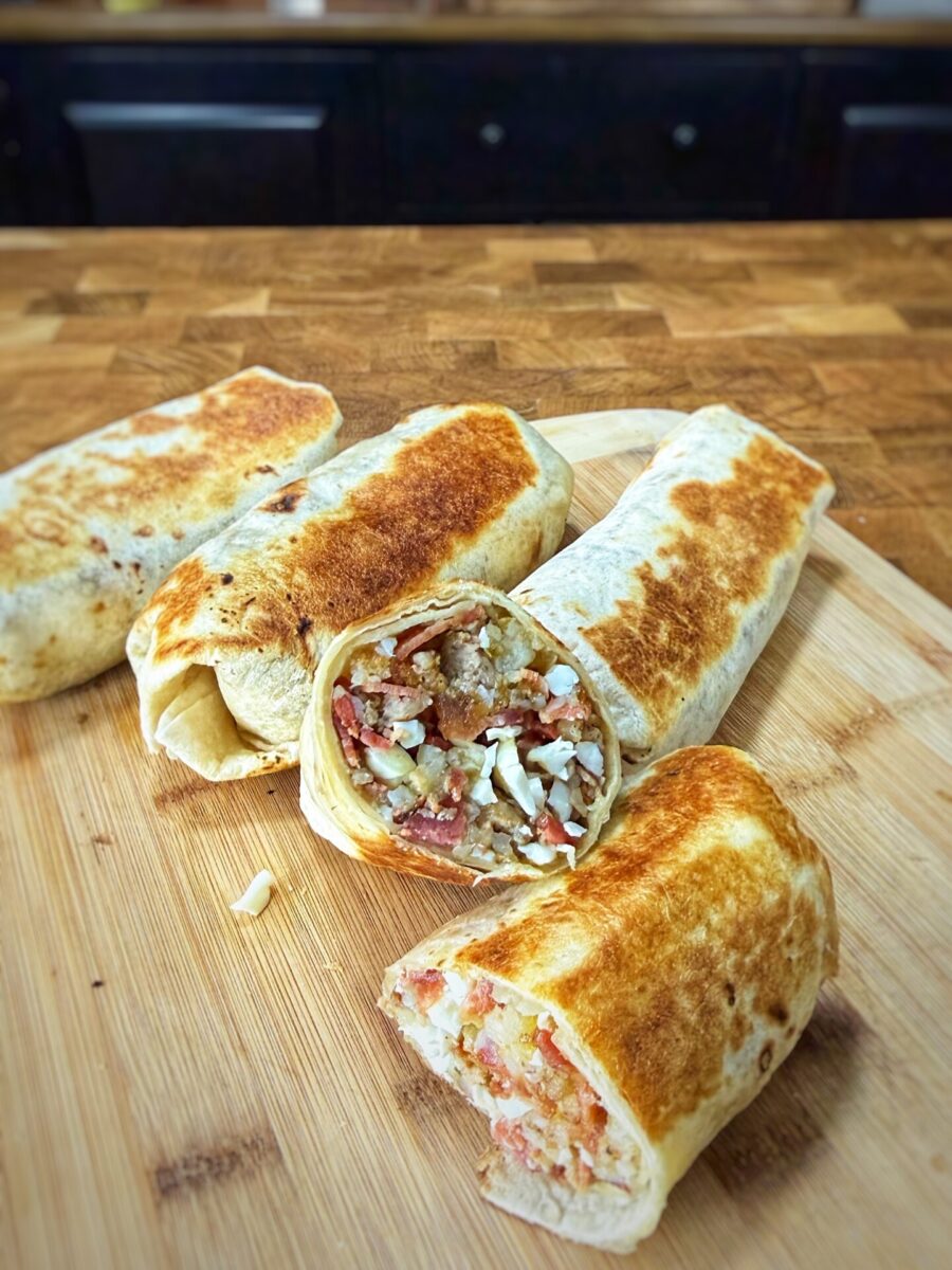 Four grilled burritos rest on a wooden cutting board, with one burrito cut open to reveal a filling of rice, meat, beans, chopped vegetables, and cheese. The background is a kitchen countertop and cabinets.