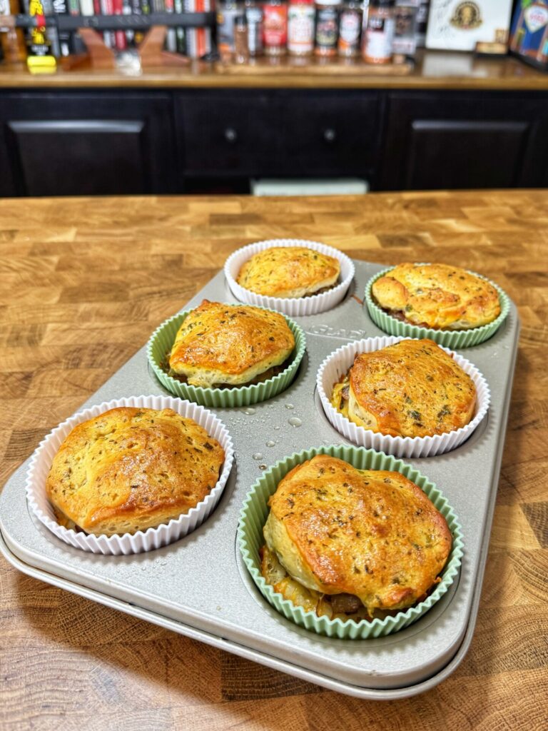 A muffin tin on a wooden countertop holds six baked savory muffins in paper liners, with a golden-brown top. Kitchen shelves with various bottles and spices are blurred in the background.
