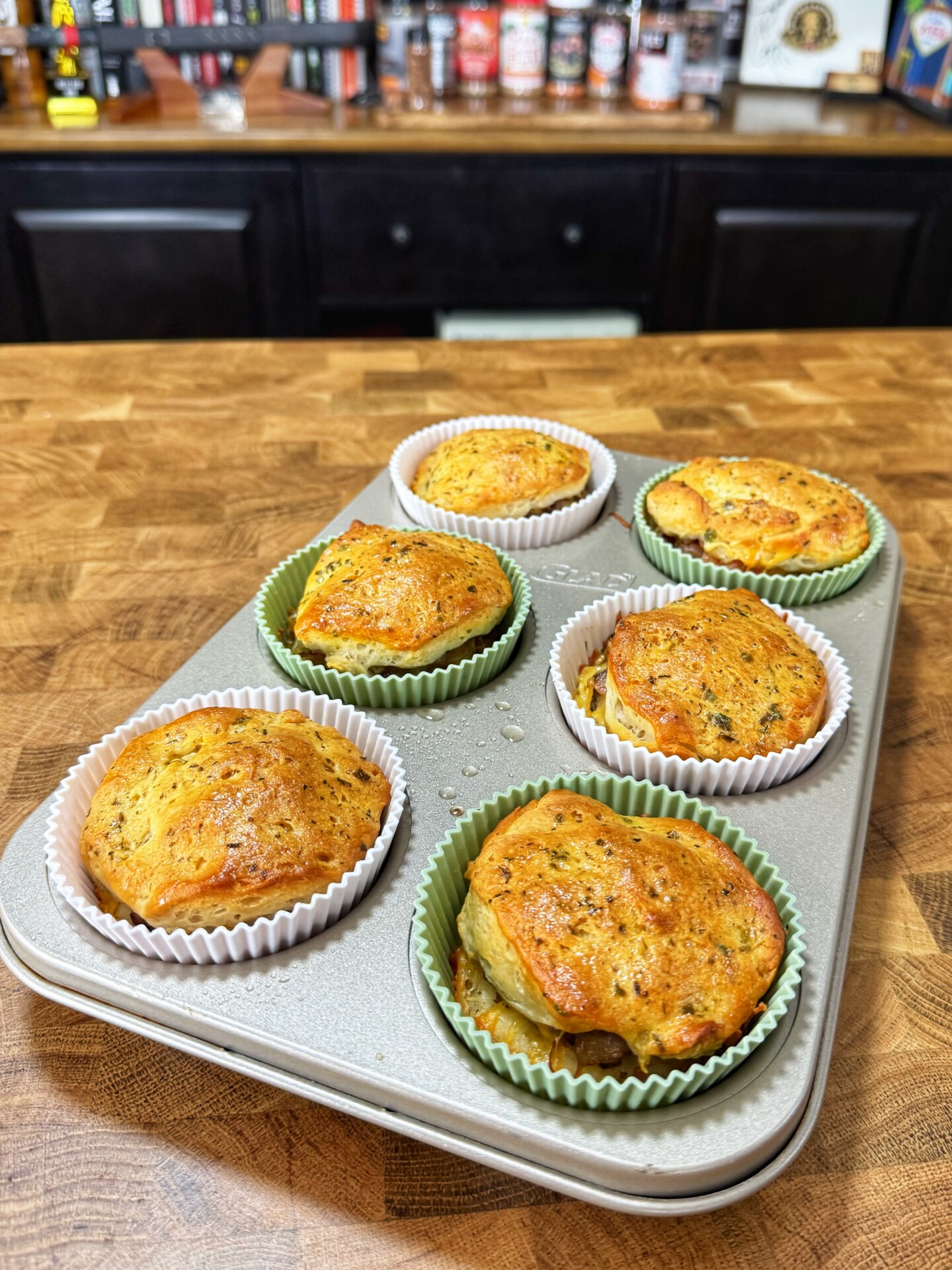A muffin tin on a wooden countertop holds six baked savory muffins in paper liners, with a golden-brown top. Kitchen shelves with various bottles and spices are blurred in the background.