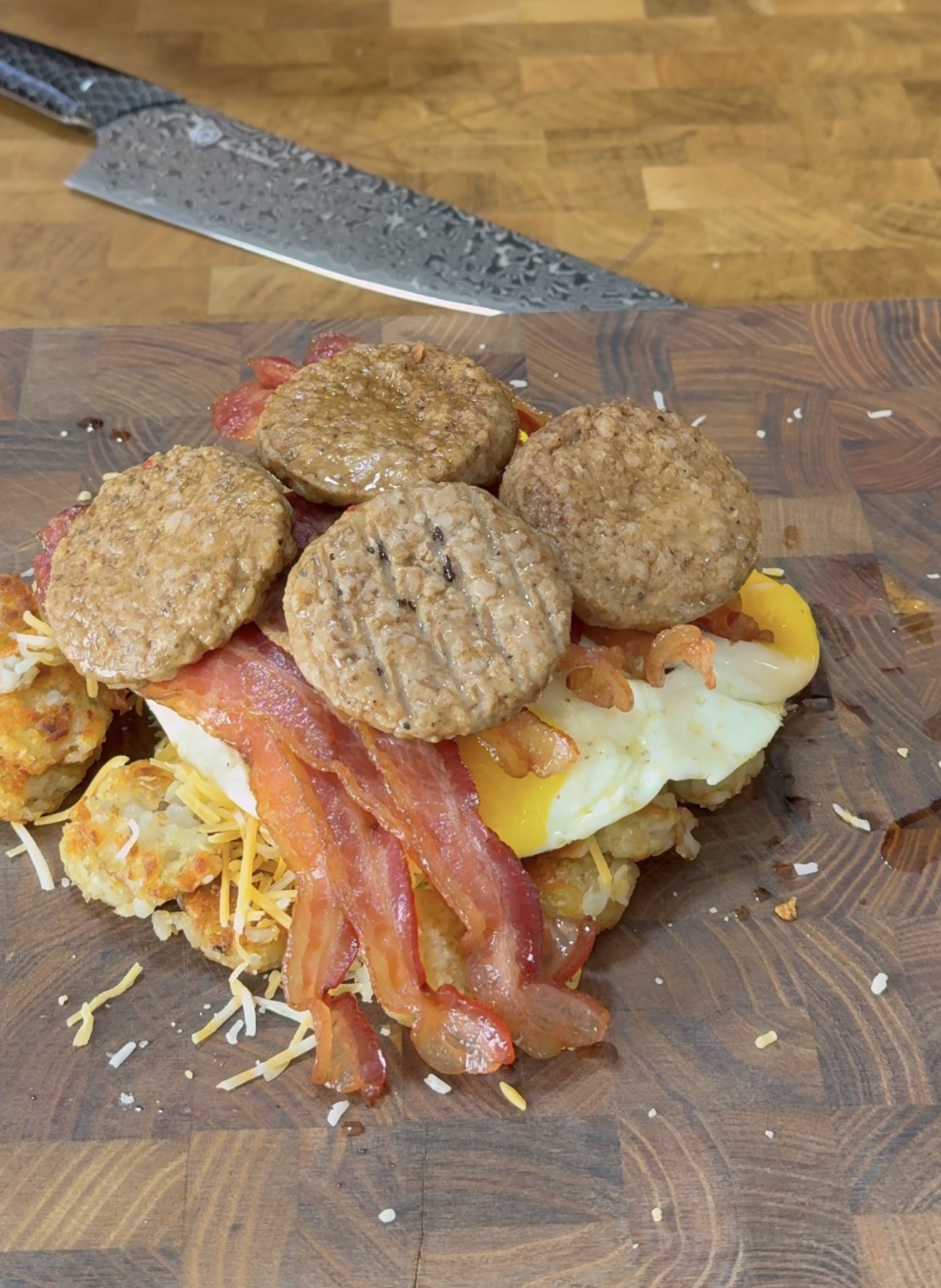 A breakfast dish on a wooden cutting board features hash browns, fried eggs, shredded cheese, strips of cooked bacon, and several round sausage patties. A knife rests in the background.