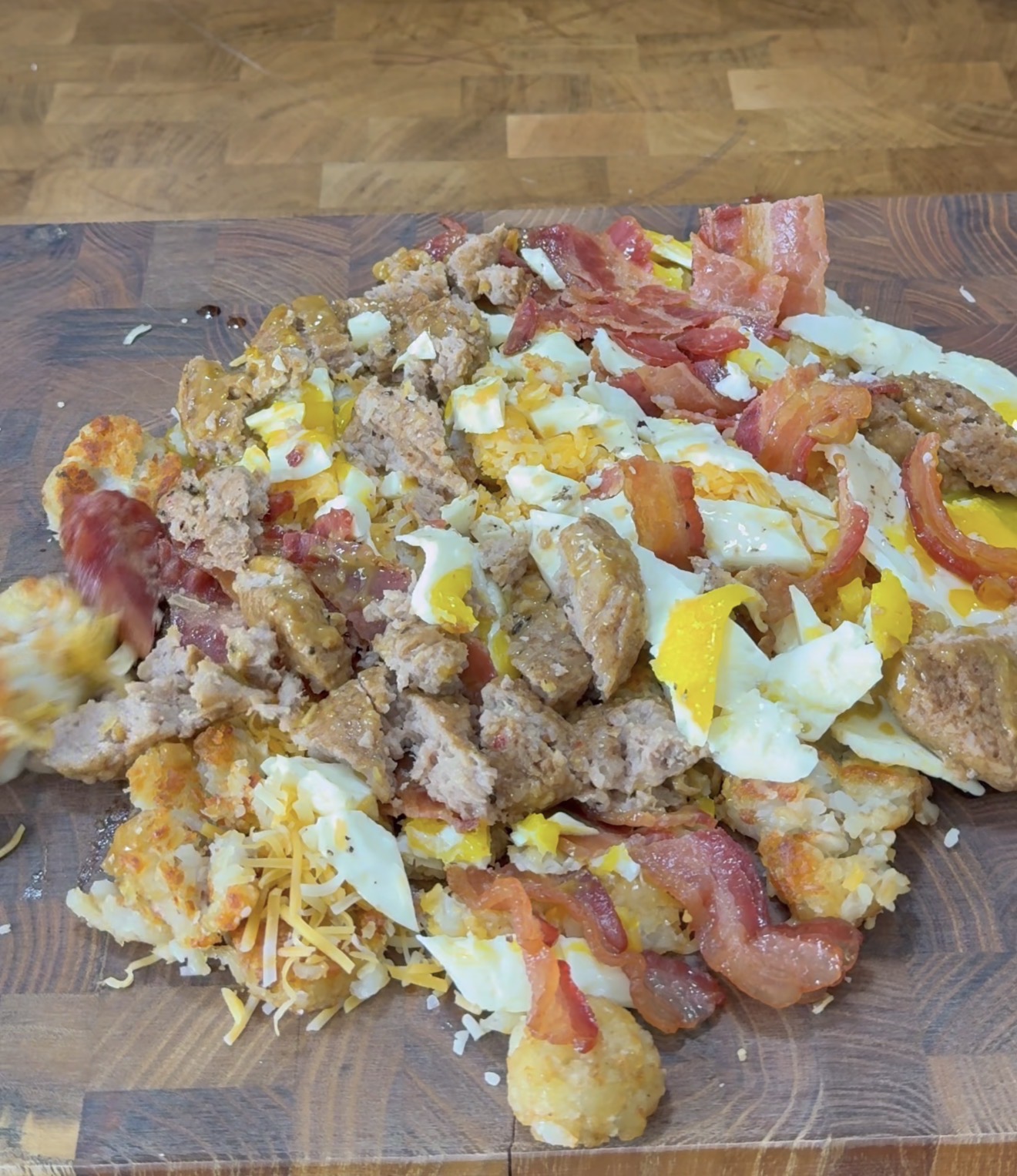 A wooden cutting board topped with a messy pile of breakfast foods including scrambled eggs, bacon strips, sausage pieces, shredded cheese, and tater tots.