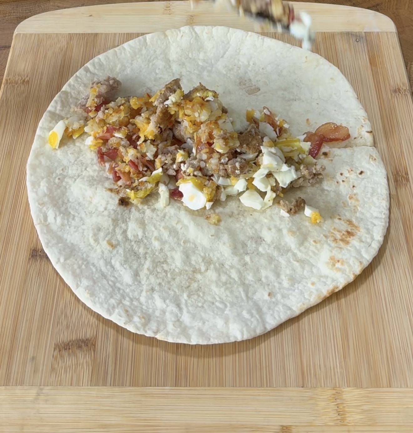 A flour tortilla on a wooden cutting board is topped with scrambled eggs, sausage, bacon, and shredded cheese, being prepared for a breakfast burrito.