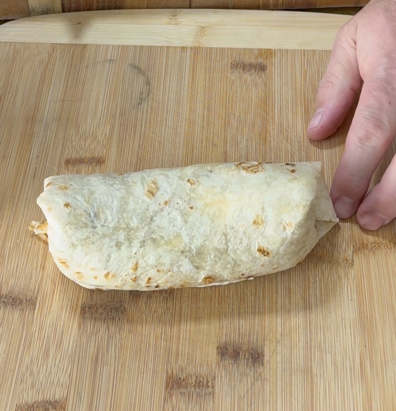 A hand holds a wrapped burrito on a wooden cutting board, ready to be served or cut.