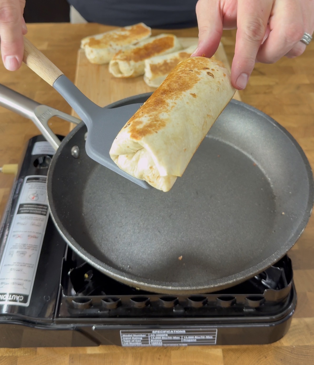 A person uses a spatula to lift a golden-brown burrito from a frying pan on a portable stove, with more burritos visible on a wooden surface in the background.