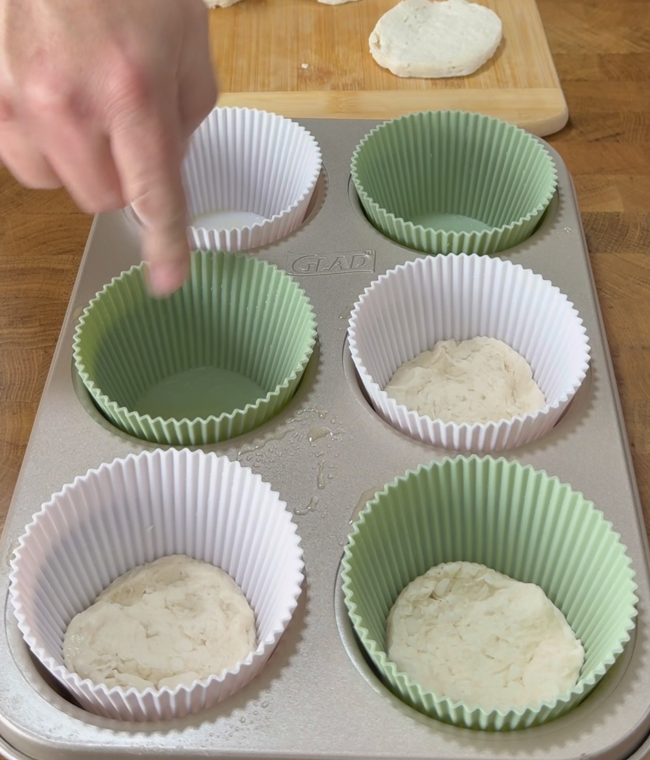 A hand places dough rounds into green and white silicone cupcake liners in a muffin tray, with more dough and a wooden cutting board visible in the background.