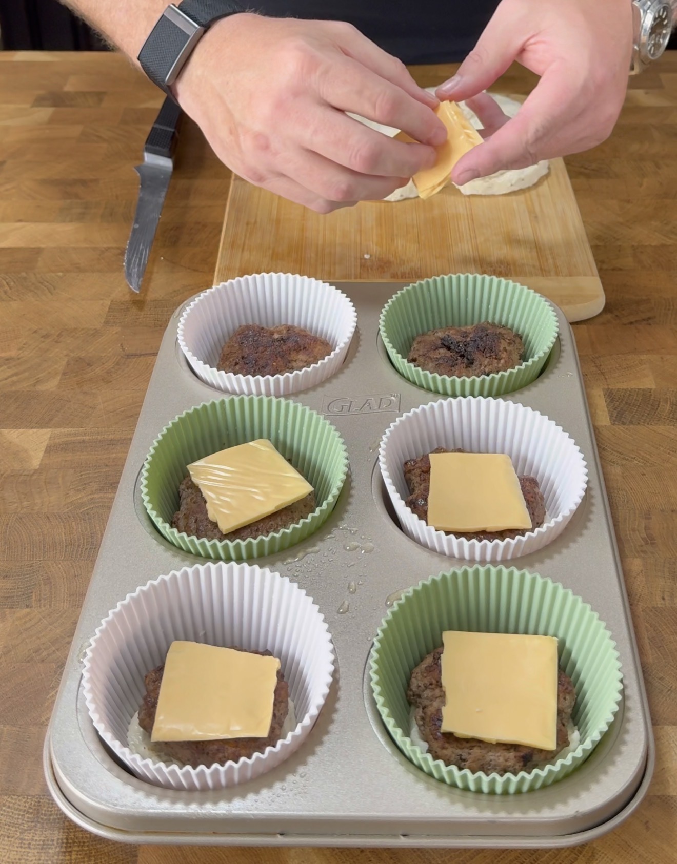 A person places slices of cheese on top of cooked burger patties in cupcake liners within a muffin tin. A cutting board and a knife are in the background on a wooden surface.