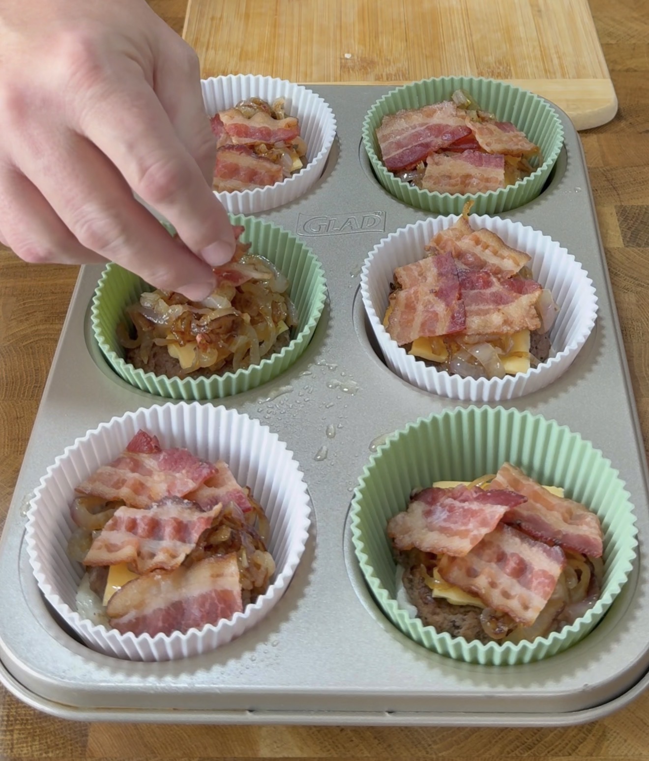 A hand sprinkles salt onto uncooked muffin cups filled with layers of caramelized onions, cheese, and strips of bacon in a metal muffin tin lined with silicone baking cups.