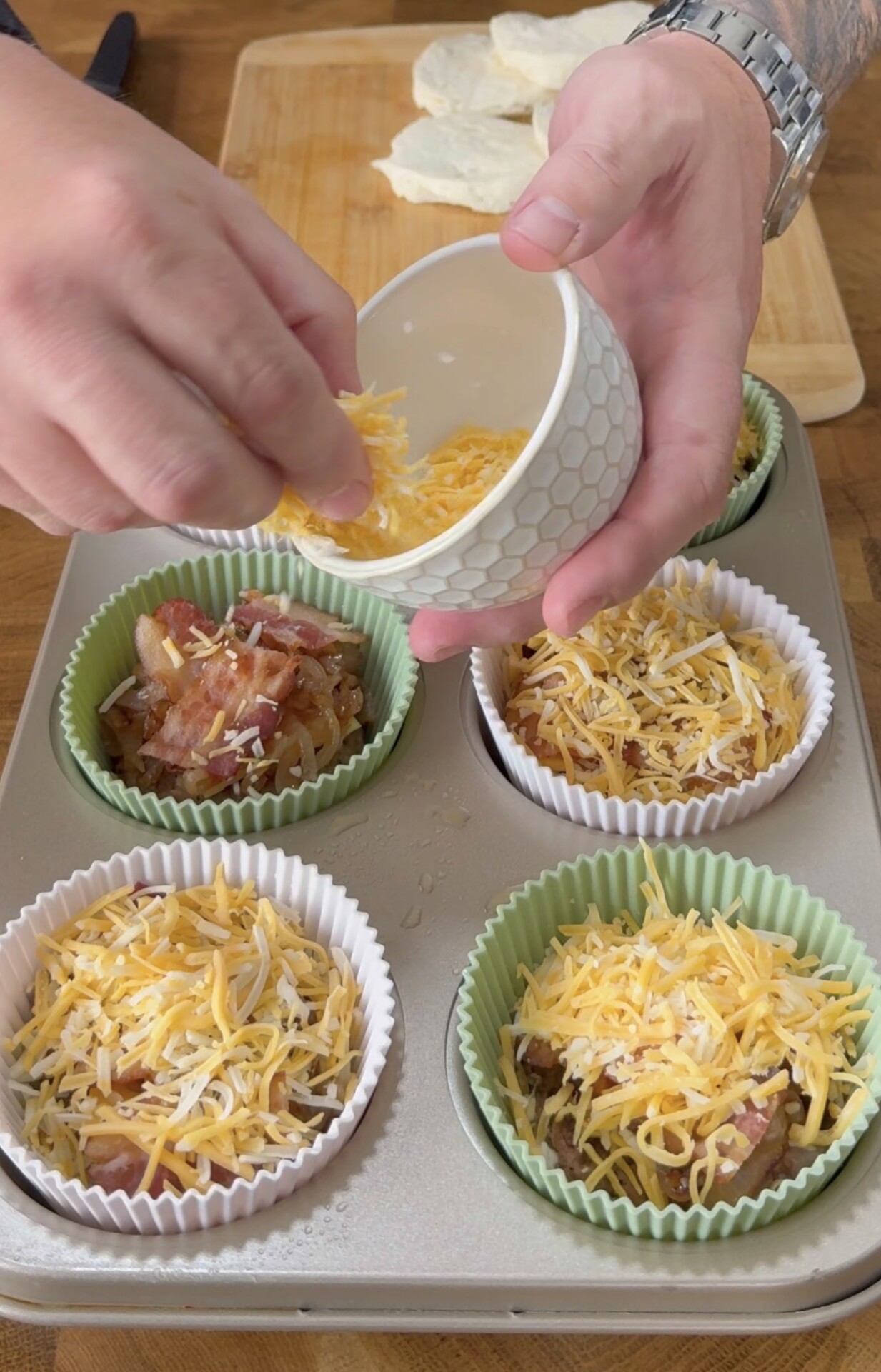 A person sprinkles shredded cheese over muffin tin cups lined with green and white wrappers, filled with bacon and other ingredients, on a wooden surface with biscuit dough visible in the background.