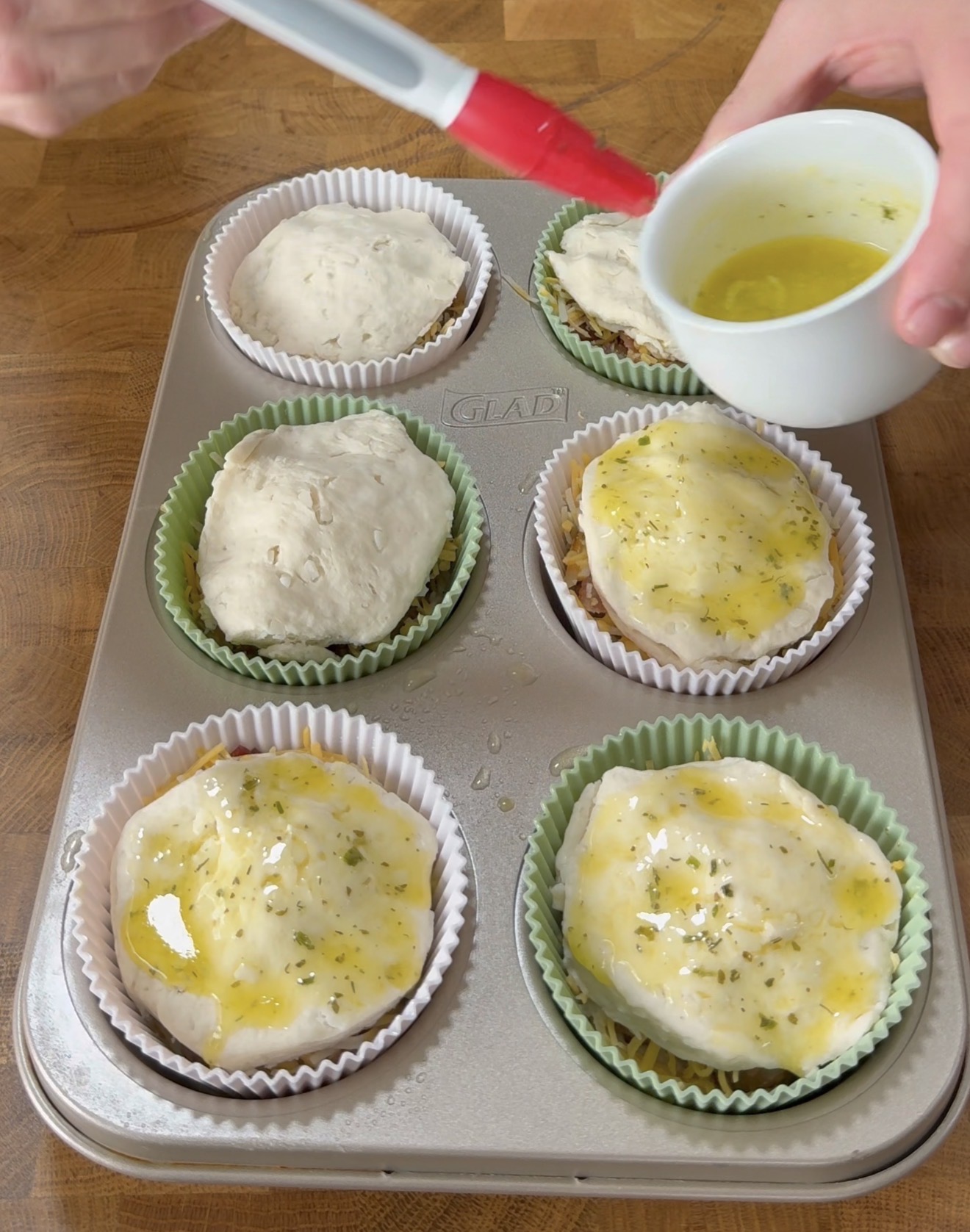 A hand brushes melted butter and herbs onto uncooked biscuit dough in a six-cup muffin tin lined with colorful paper liners on a wooden surface.