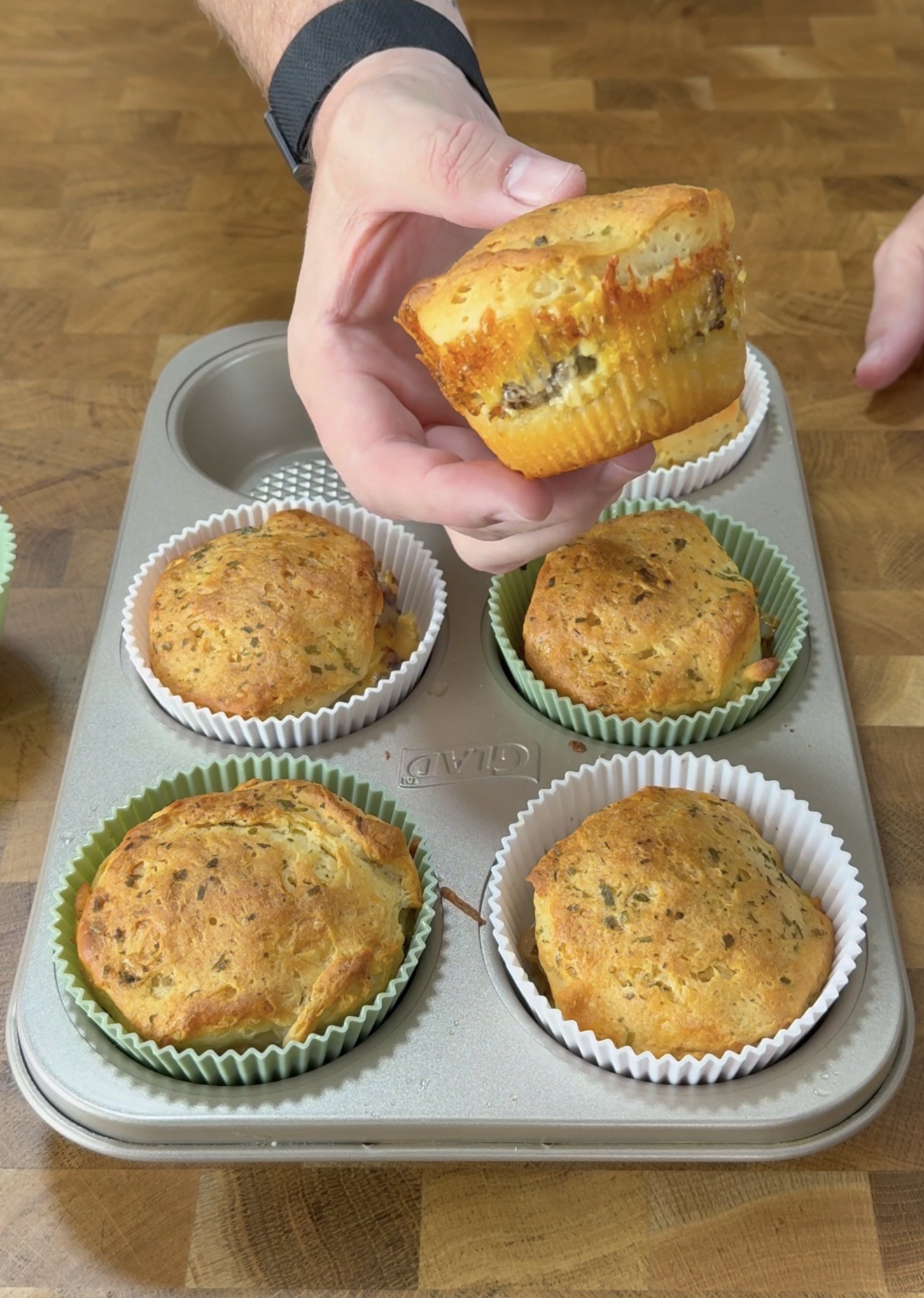 A hand holds a savory muffin with a visible filling over a metal muffin tray containing four other baked muffins in green and white paper liners on a wooden surface.