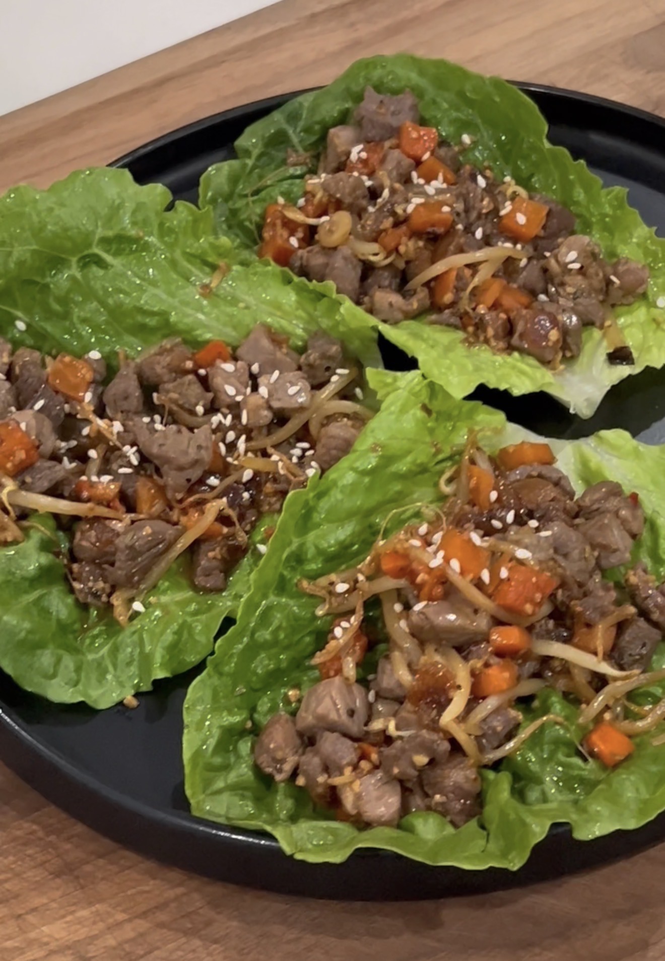 Three lettuce leaves topped with diced beef, shredded carrots, bean sprouts, and sprinkled with sesame seeds are arranged on a black plate on a wooden surface.