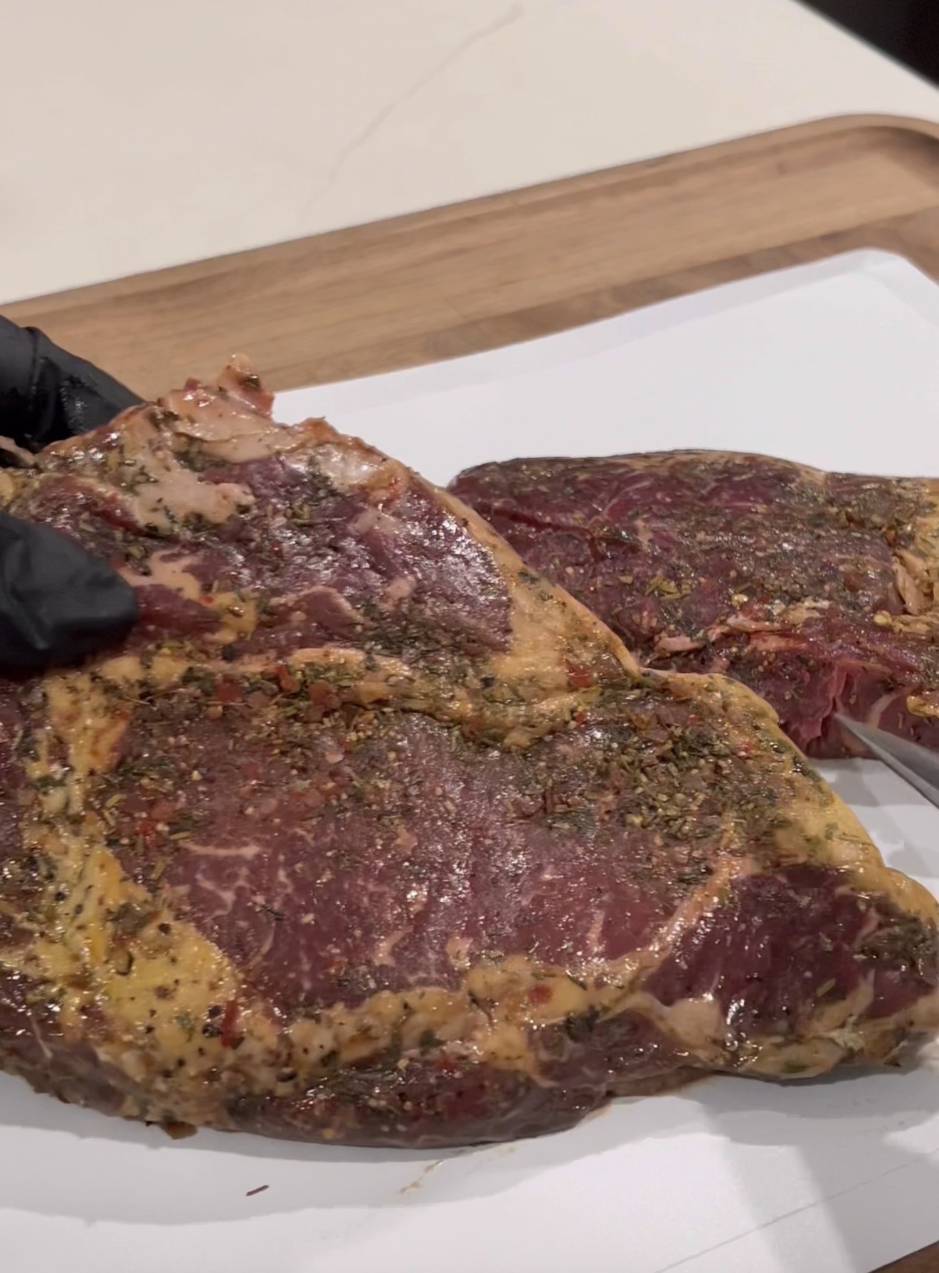 A gloved hand holds a seasoned raw steak over a white surface, with another piece of seasoned raw steak resting nearby on the same surface. A wooden cutting board is visible in the background.