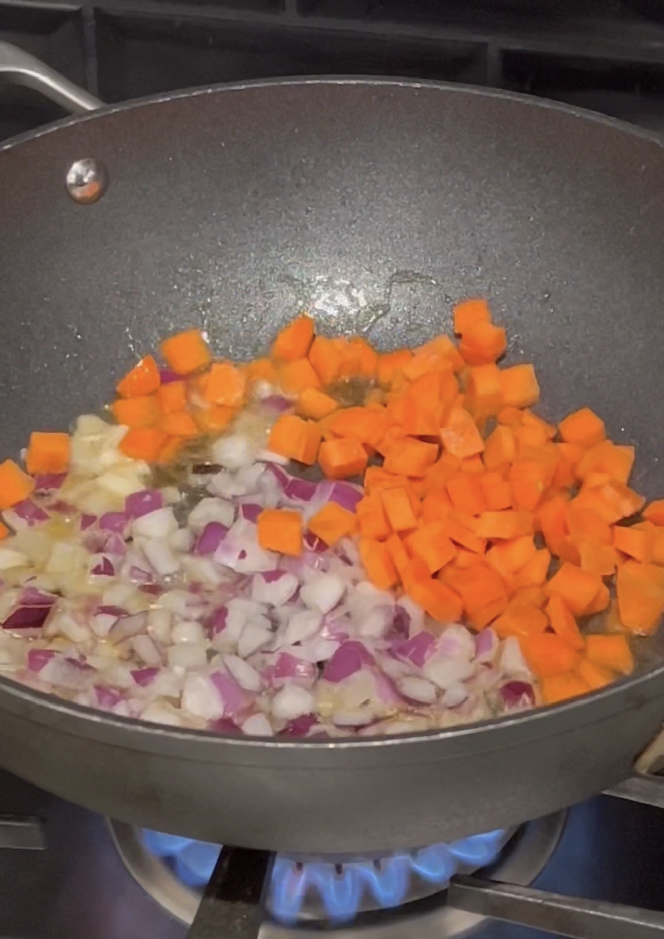 A wok on a stove with chopped red onions and diced carrots sautéing in oil over a blue flame.