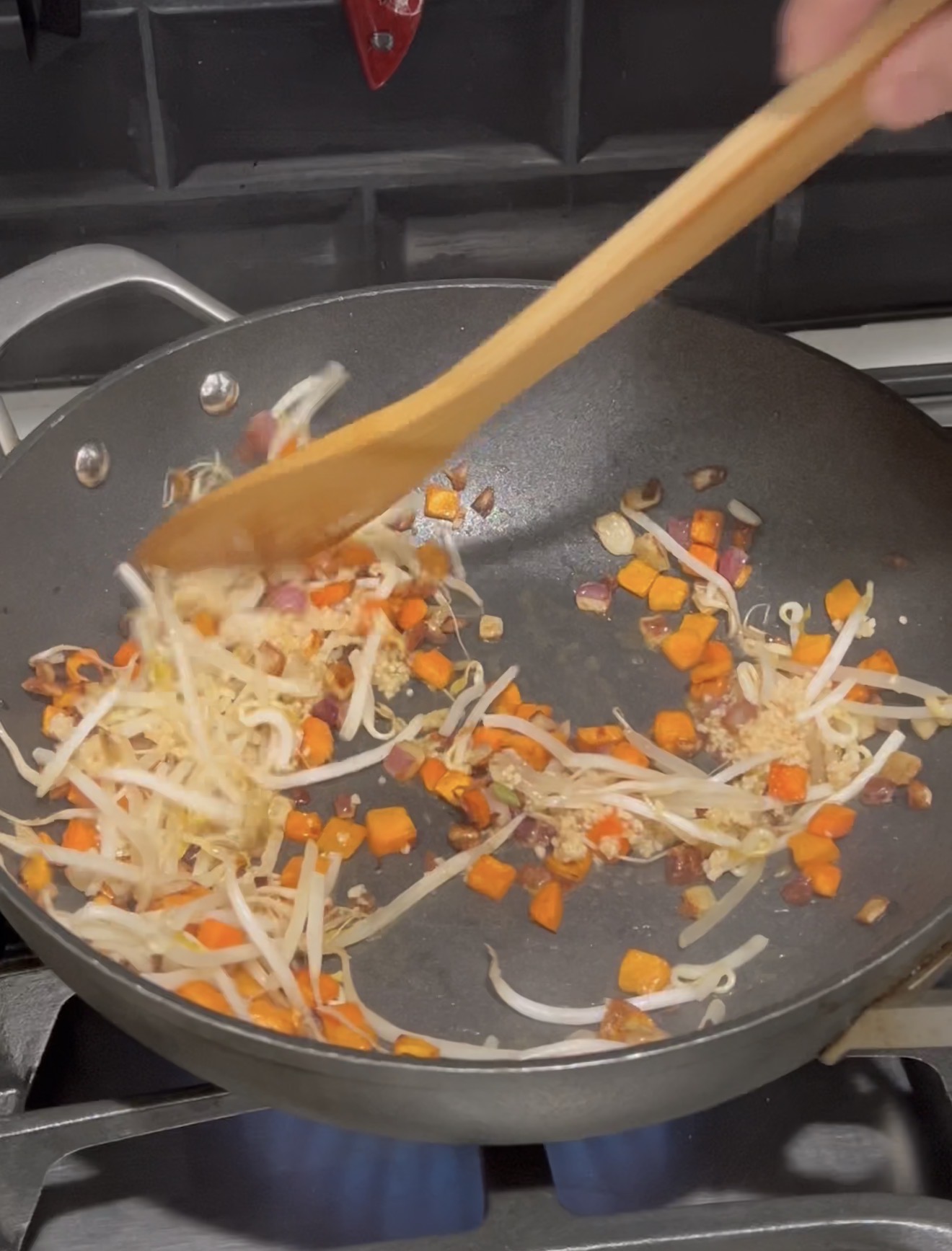 A hand stirs chopped carrots, onions, and bean sprouts in a black wok on a gas stove with a wooden spatula.