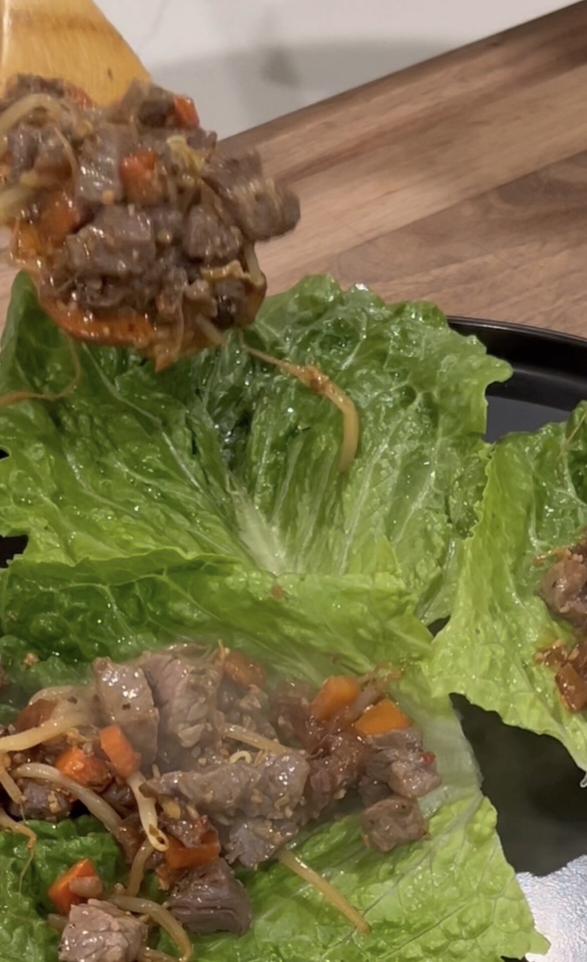 A wooden spoon holds a portion of cooked beef, carrots, and bean sprouts above fresh lettuce leaves filled with the same mixture, ready to be wrapped and eaten.