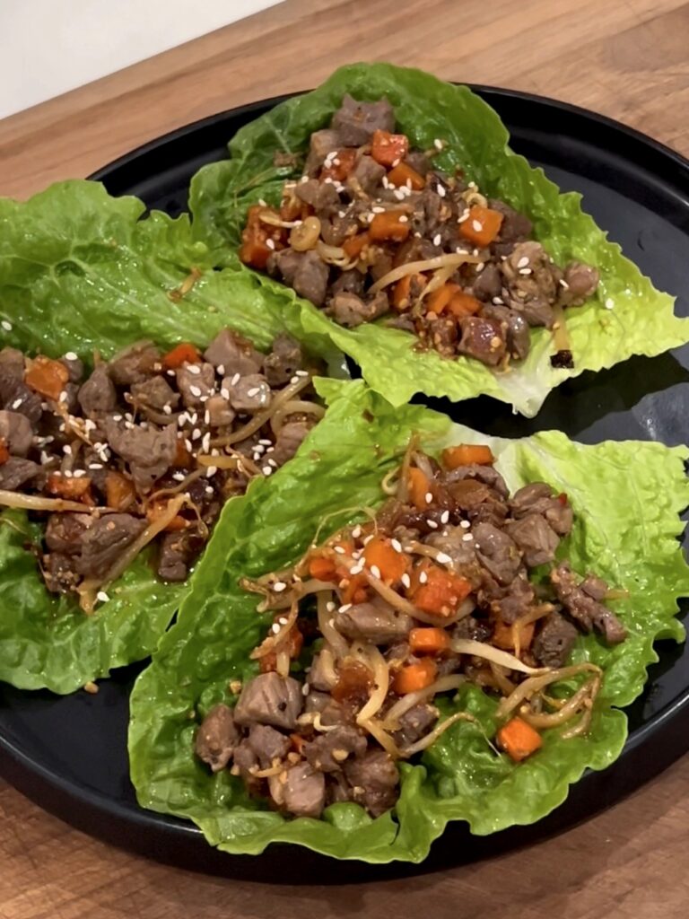 Three lettuce leaves filled with chopped beef, diced carrots, onions, and topped with sesame seeds are arranged on a black plate atop a wooden surface.